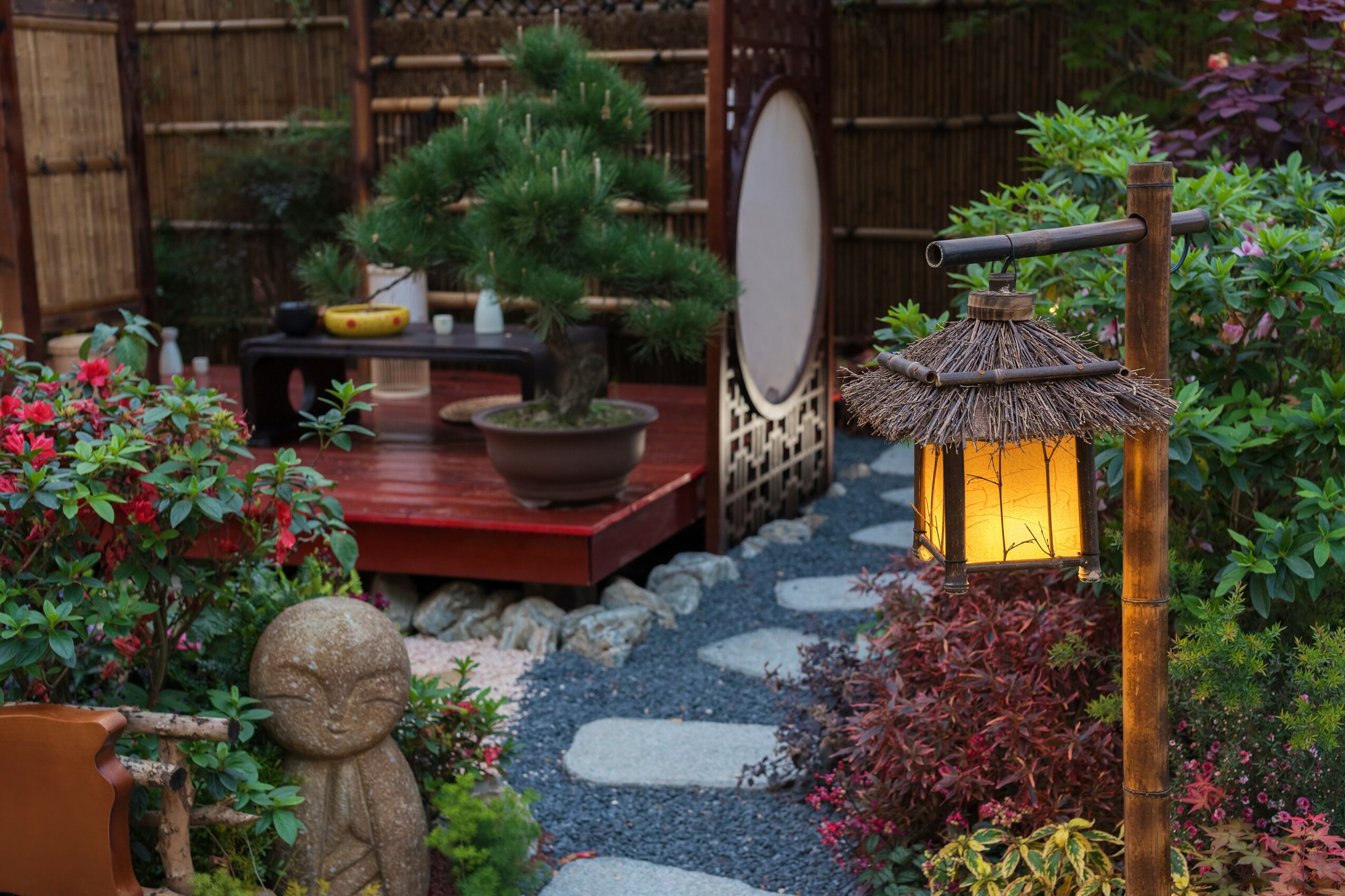 Garden scene with a stone pathway, lantern, bonsai tree, and colorful plants, enclosed by a wooden fence.