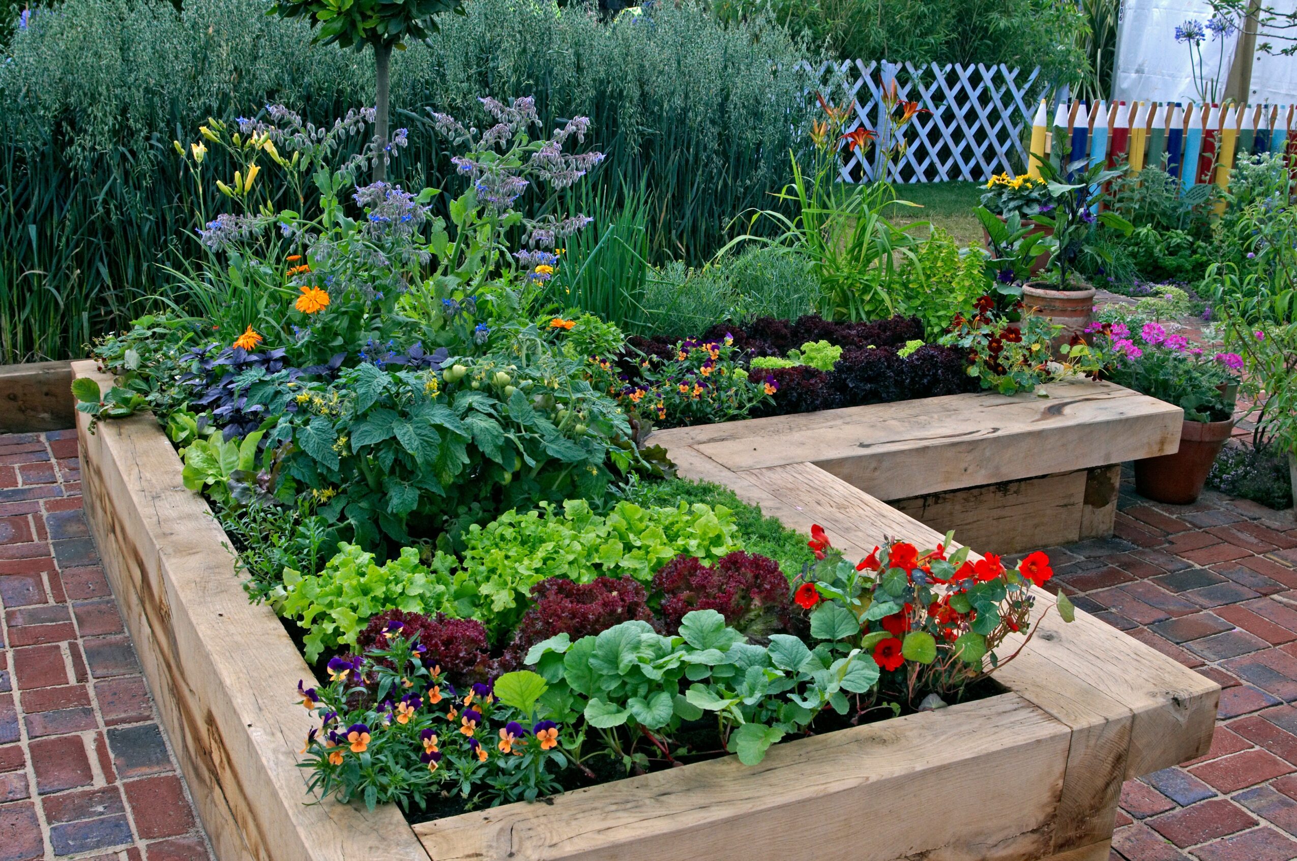Raised garden bed with colorful flowers and green plants on brick patio, background of trees and a white fence.