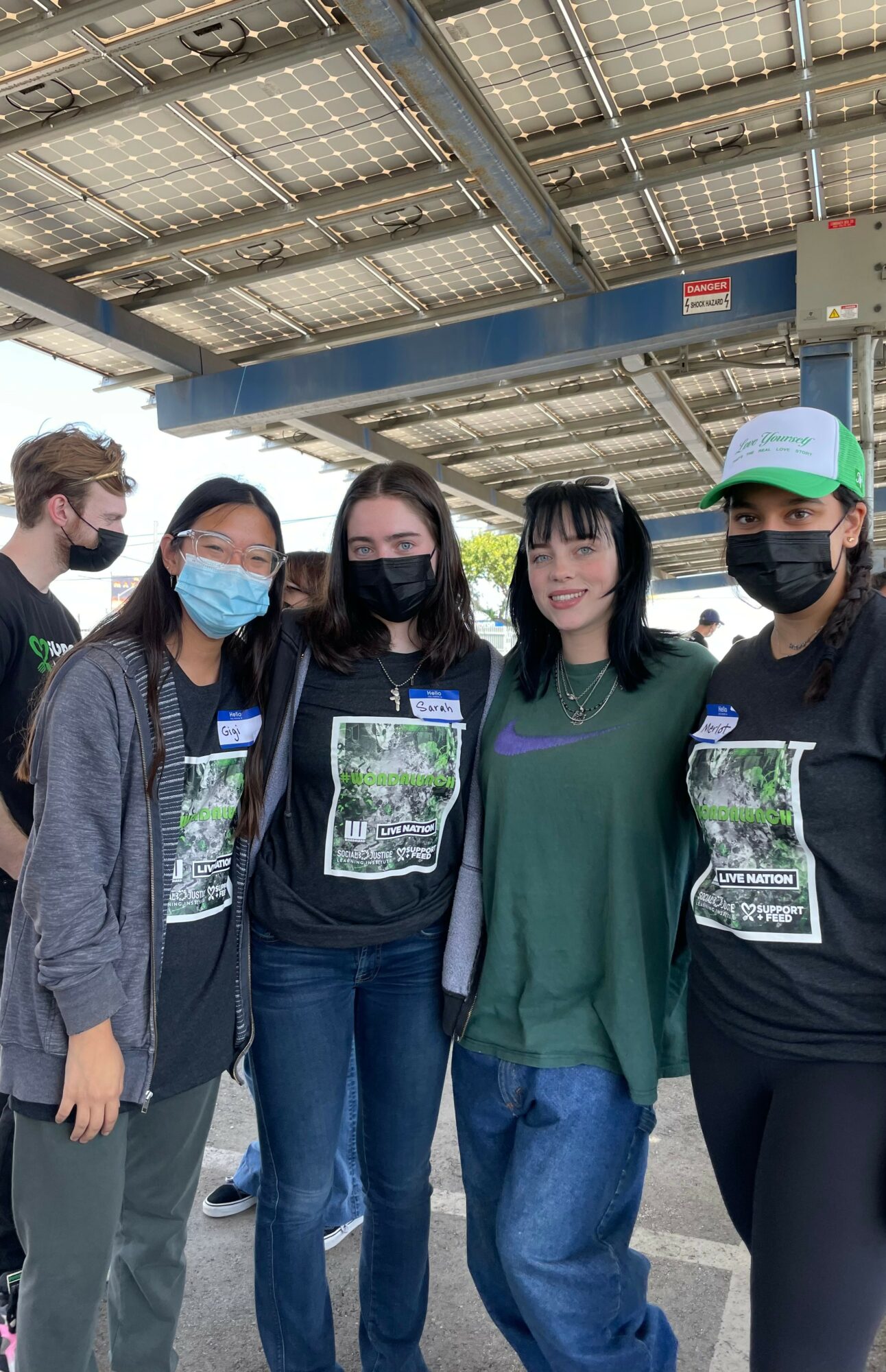 Group of five people wearing masks standing outdoors under a solar panel structure, smiling at the camera.