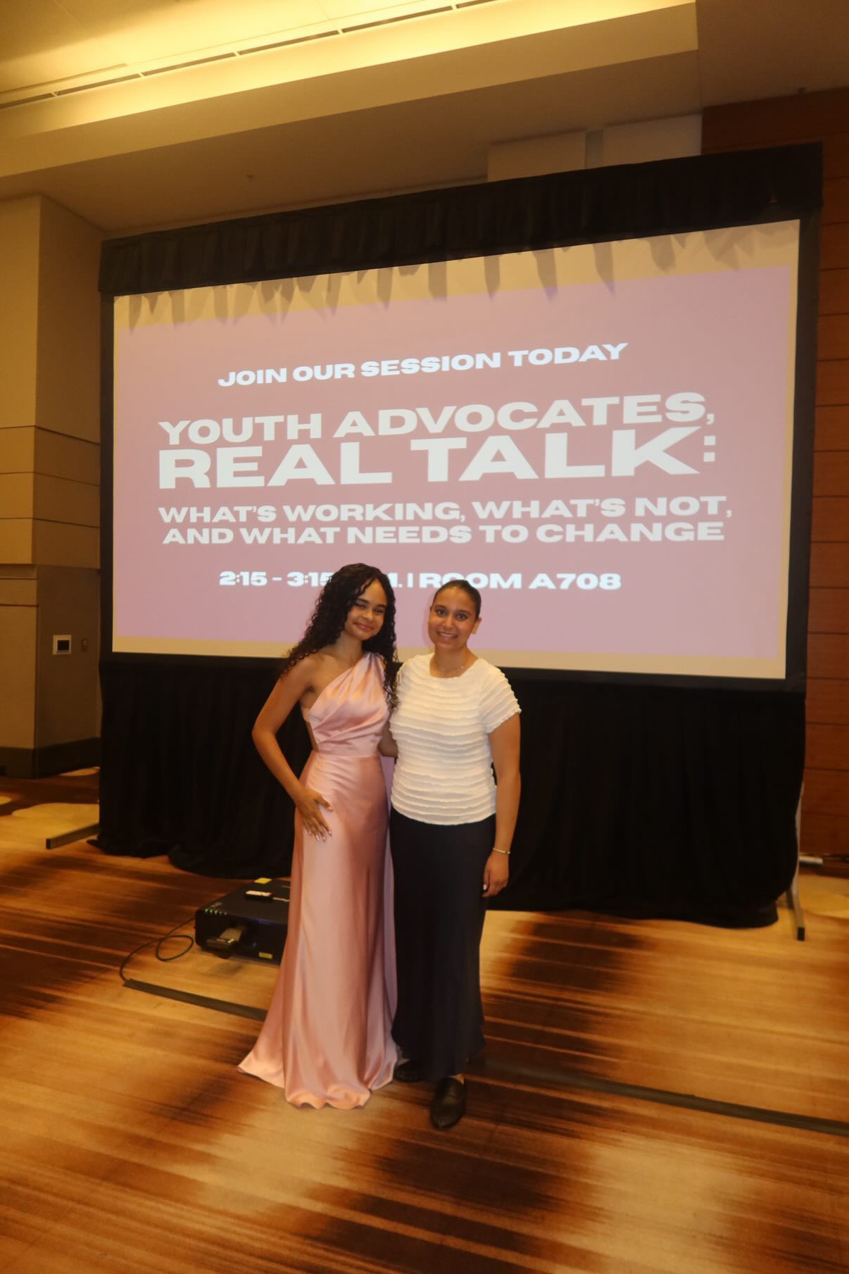 Two women stand in front of a large screen displaying event information, one in a pink gown and the other in casual attire.