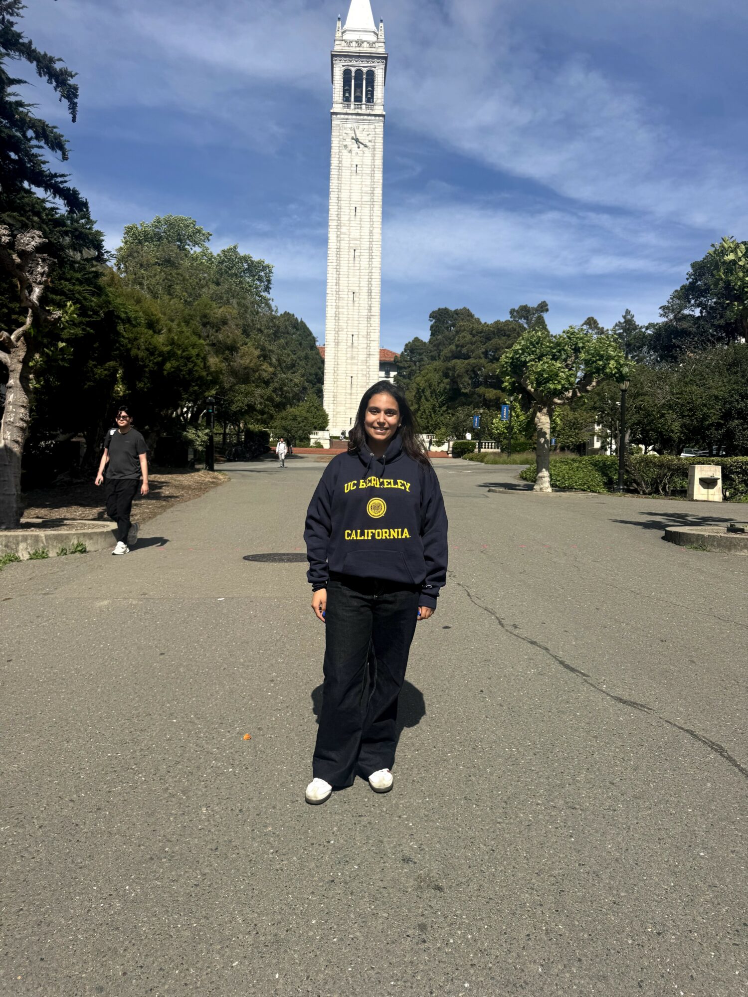 Person standing in front of a tall clock tower with trees and a blue sky, smiling, wearing a dark hoodie and pants.