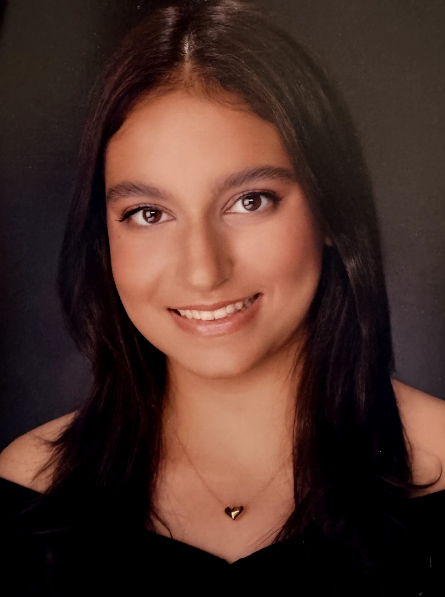 Young woman with long dark hair smiling, wearing a black top and a necklace with a heart pendant.