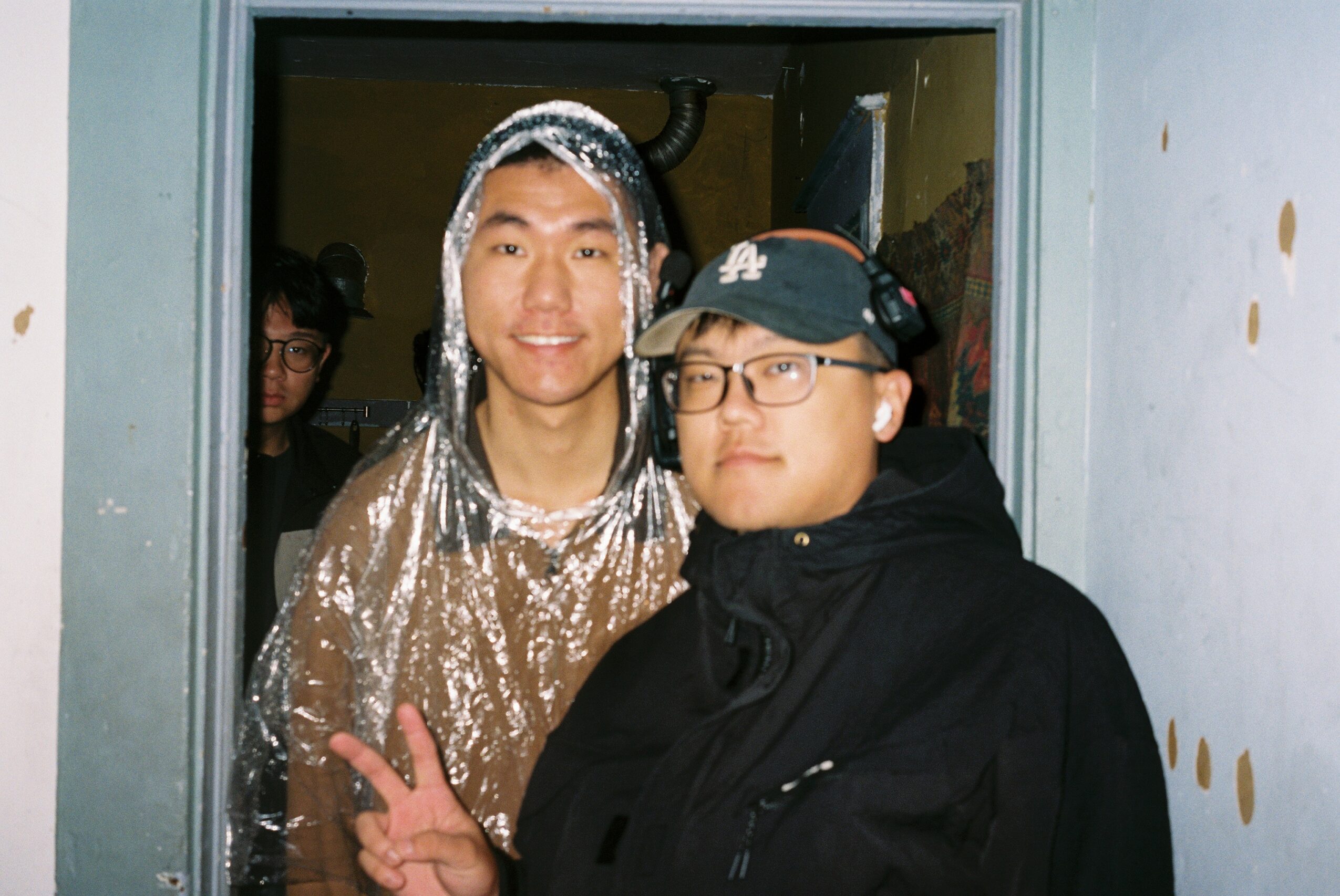 Two people standing in a doorway, one smiling and the other making a peace sign, with a person in the background.