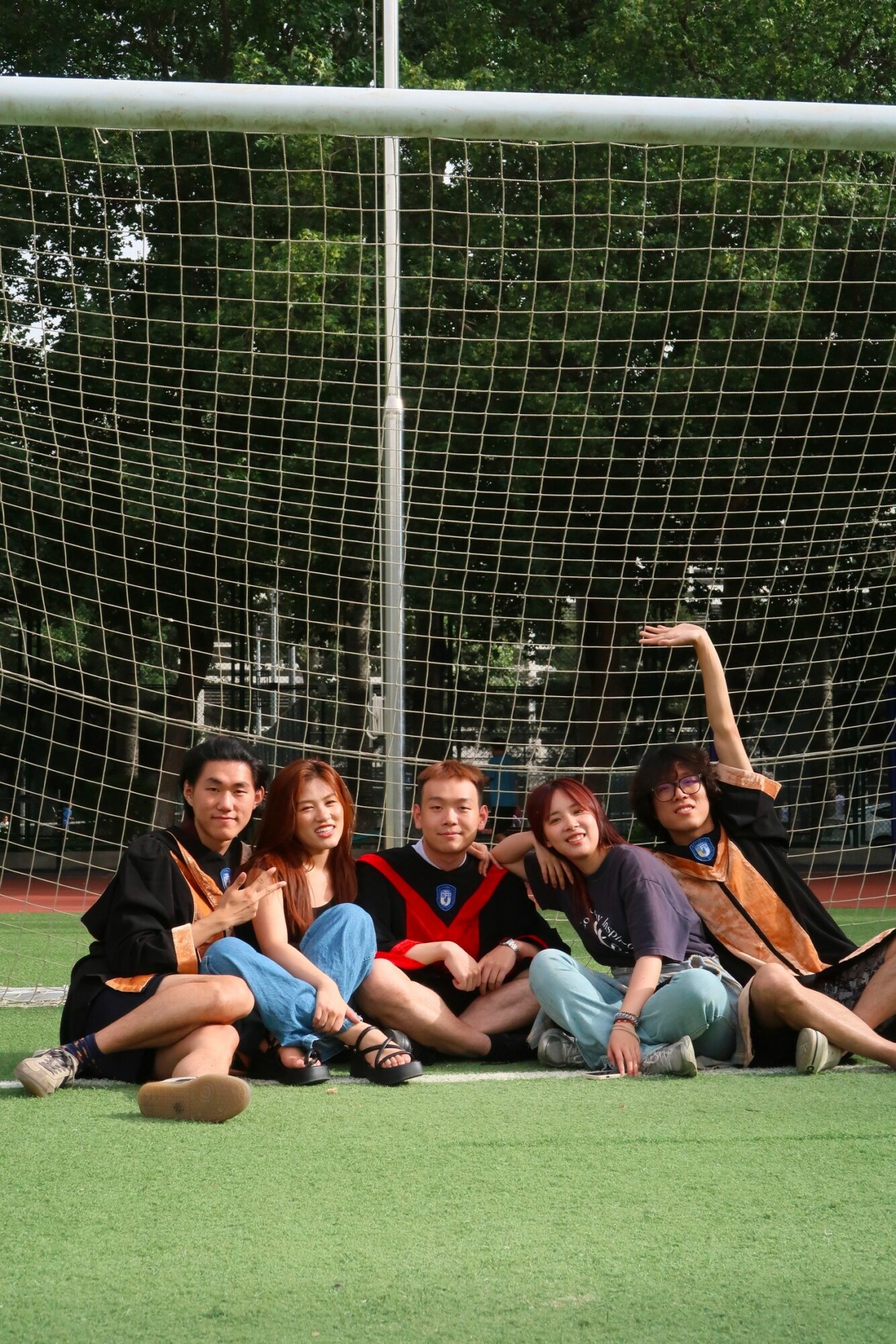 Five people sitting on a sports field in front of a net, with trees in the background.