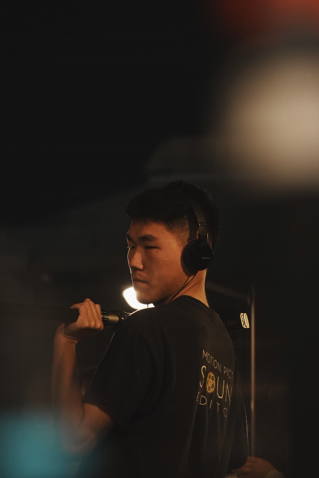 Young person with headphones holding a microphone, facing left, in a dark setting with some light behind.