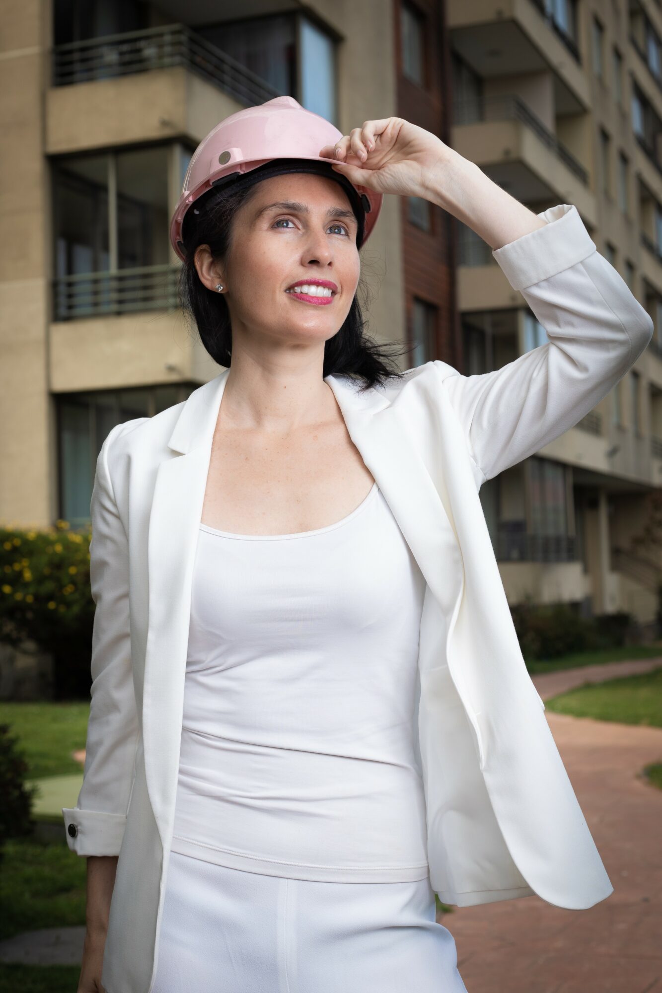 Woman wearing a pink safety helmet and white outfit, smiling outdoors in front of apartment buildings.