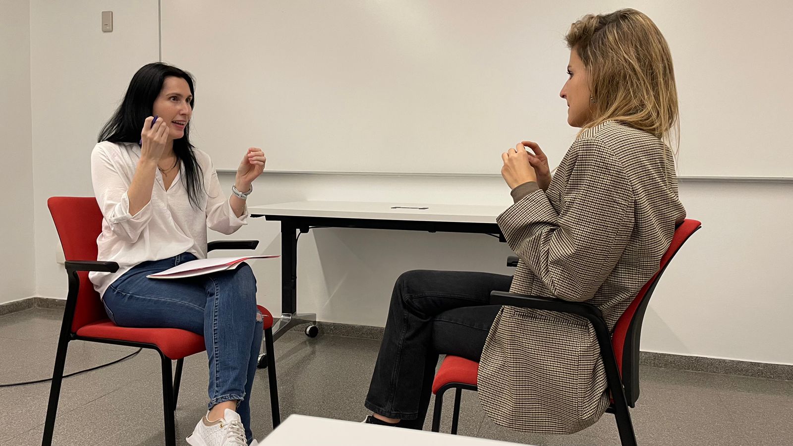 Two women sit facing each other in a room, engaged in conversation, with a whiteboard behind them.