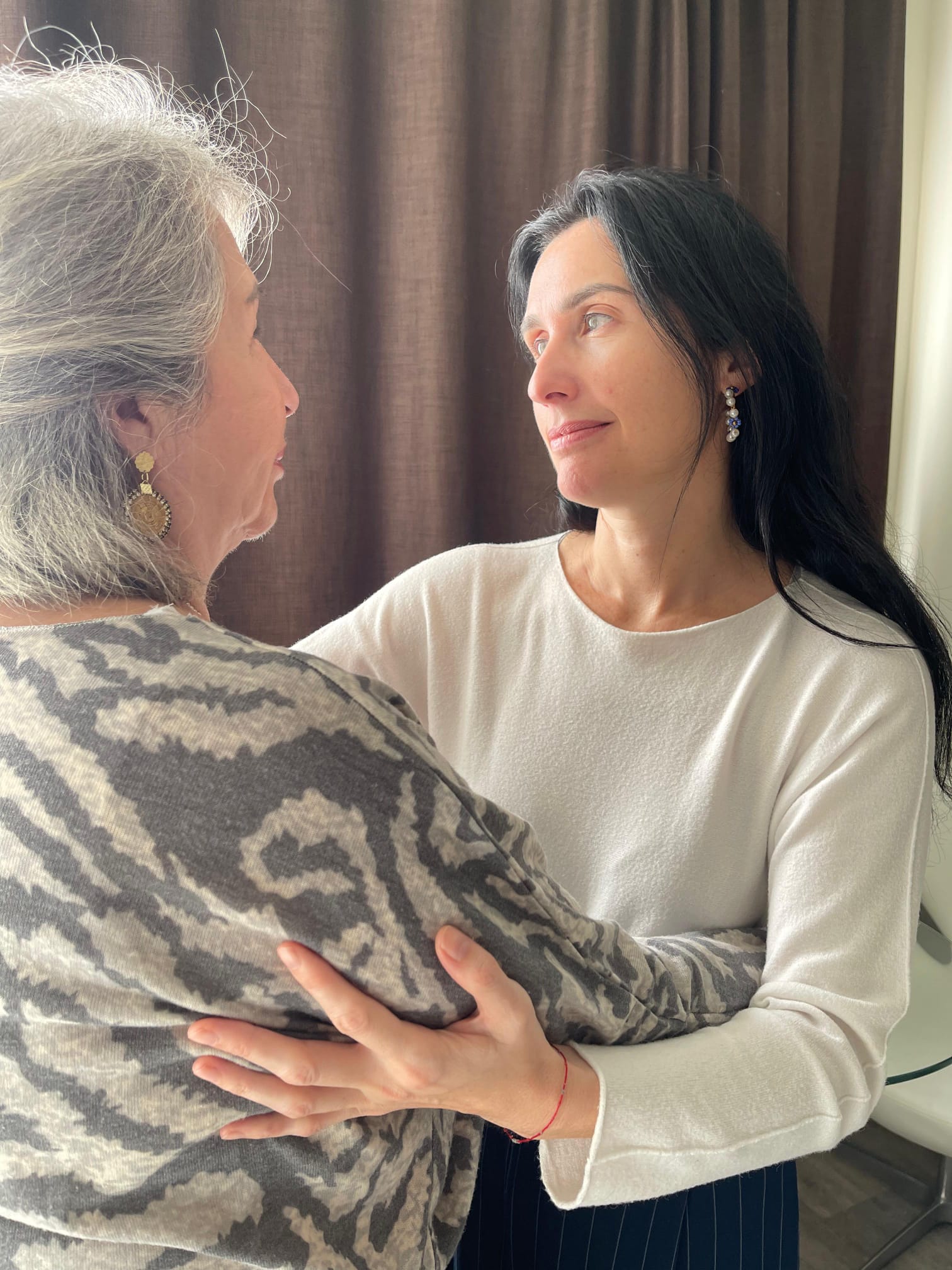 Two women face each other, one with gray hair and patterned clothing, the other with dark hair and a white top, holding hands.