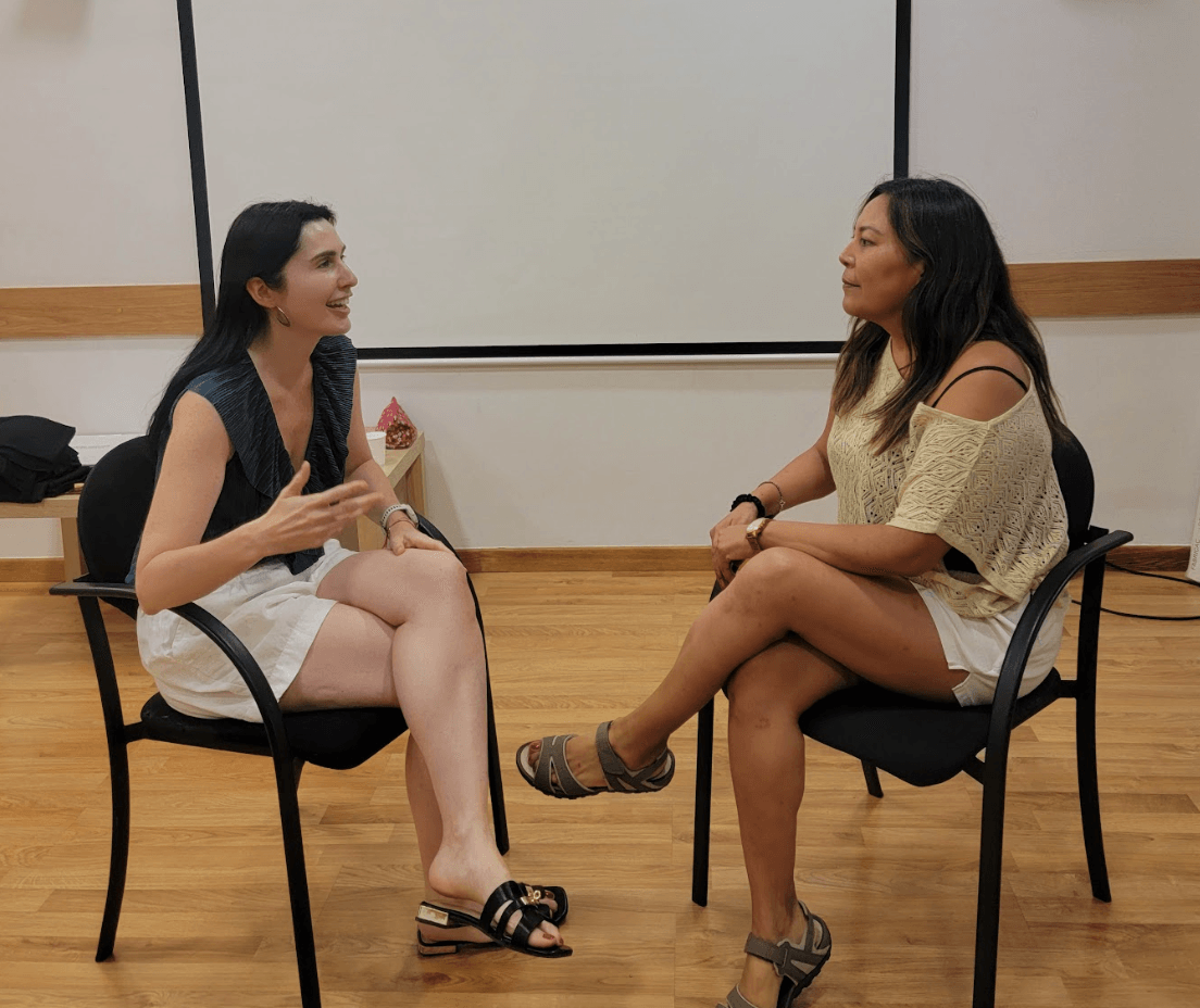 Two women sit facing each other in chairs, engaged in conversation in a room with a whiteboard and wooden floor.