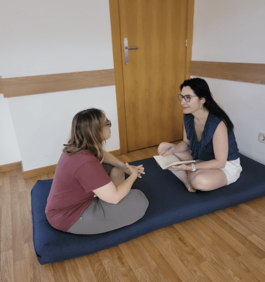Two women sitting on a blue mattress in a room, one holding a notebook, engaging in conversation.
