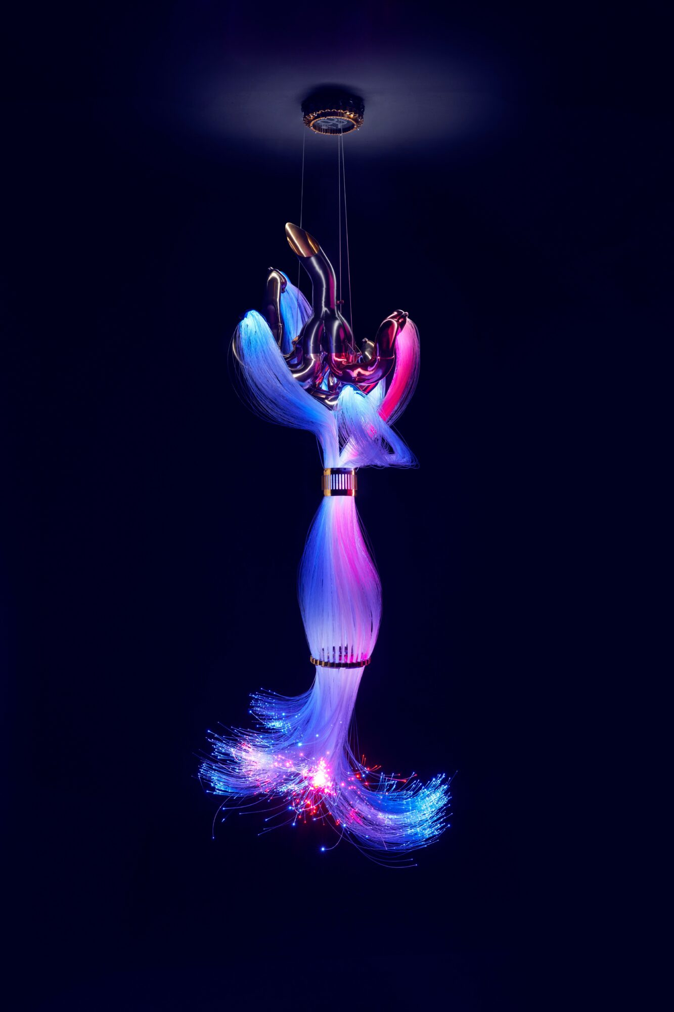Colorful, illuminated sculpture resembling a flower or abstract form, hanging from a ceiling against a dark background.