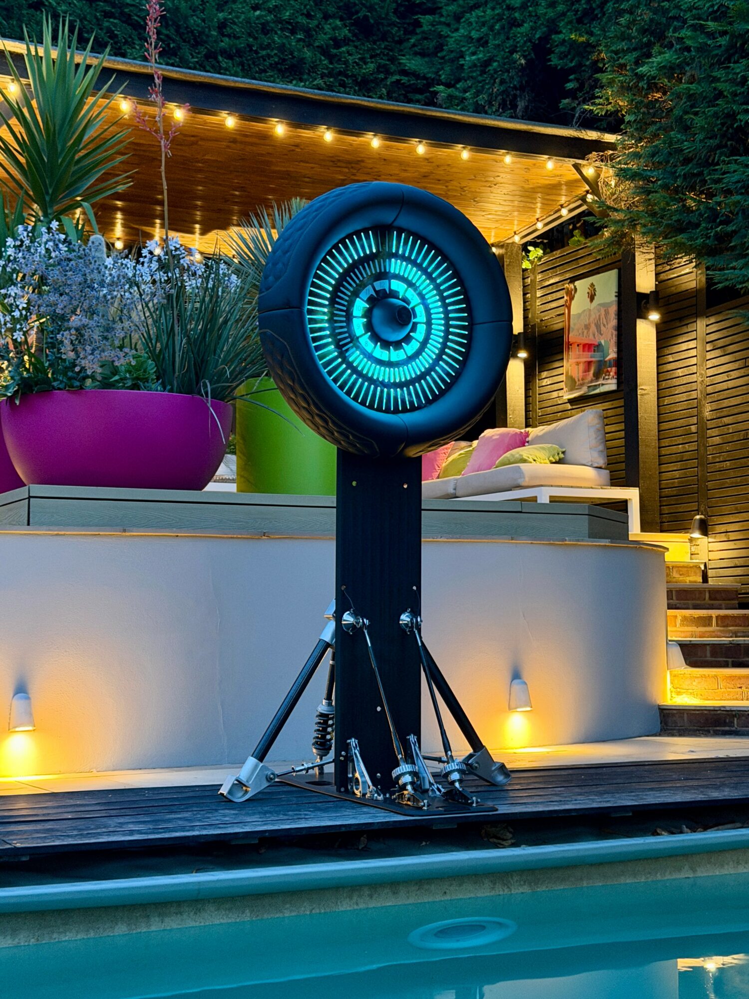 Large circular speaker or light fixture on a stand outdoors at night, with a modern patio and greenery in background.