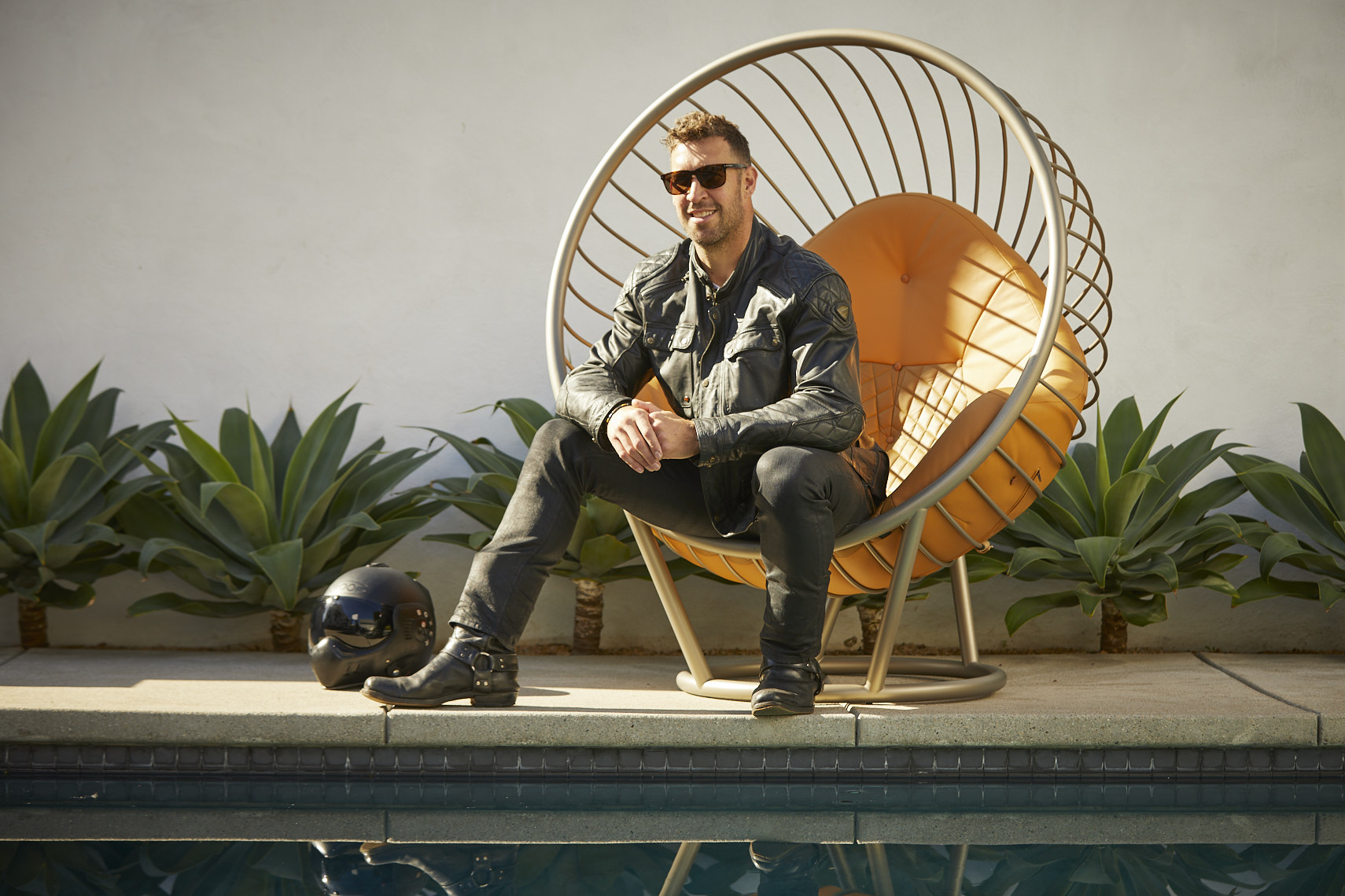 Man sitting on a modern outdoor chair with a yellow cushion, wearing sunglasses and a leather jacket, next to a pool.