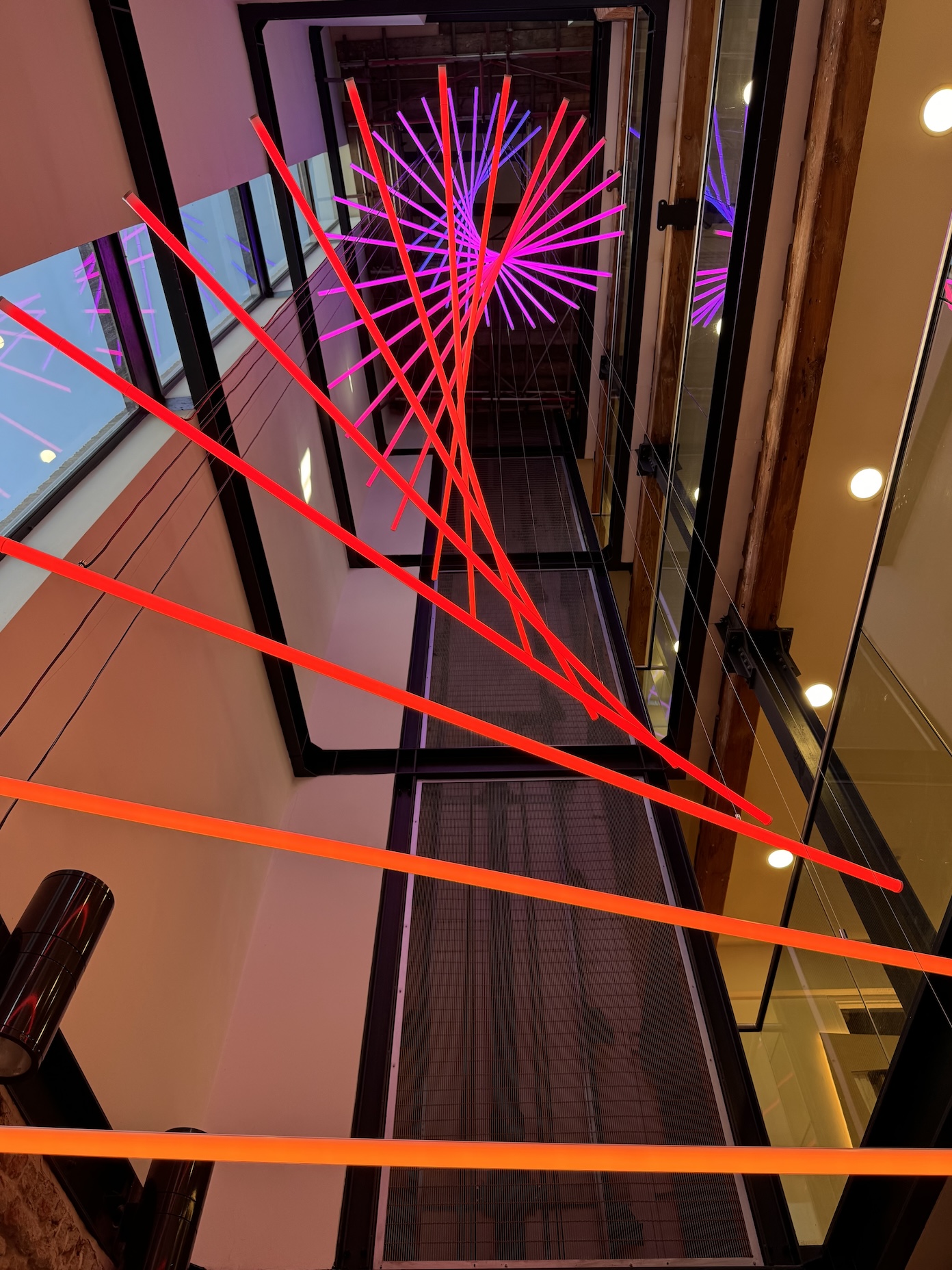 View of a glass elevator shaft with colorful laser light projections and reflections, looking downward. Word count: 20