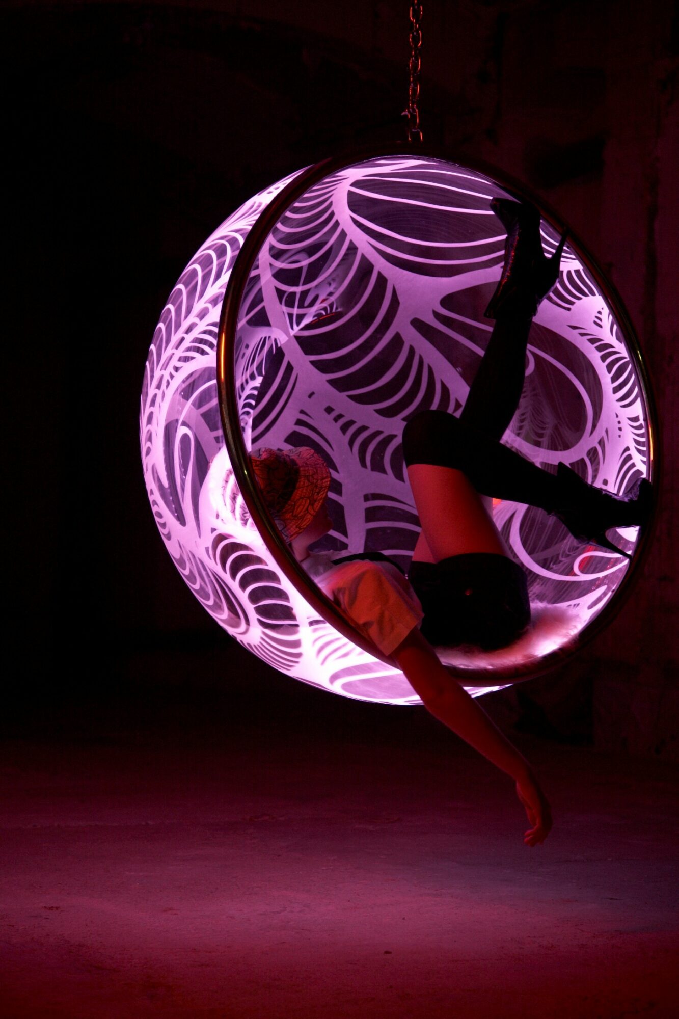 Person sitting inside a glowing, patterned spherical structure with purple and white swirling designs, hanging in a dark space.