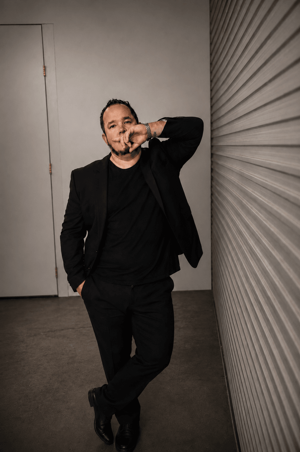 Person standing against a wall, wearing a black jacket and pants, with one hand in pocket and the other near face.