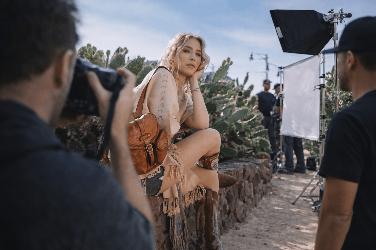 A woman with blonde hair poses outdoors during a photoshoot, with a photographer and assistant nearby, and a cactus garden in the background.