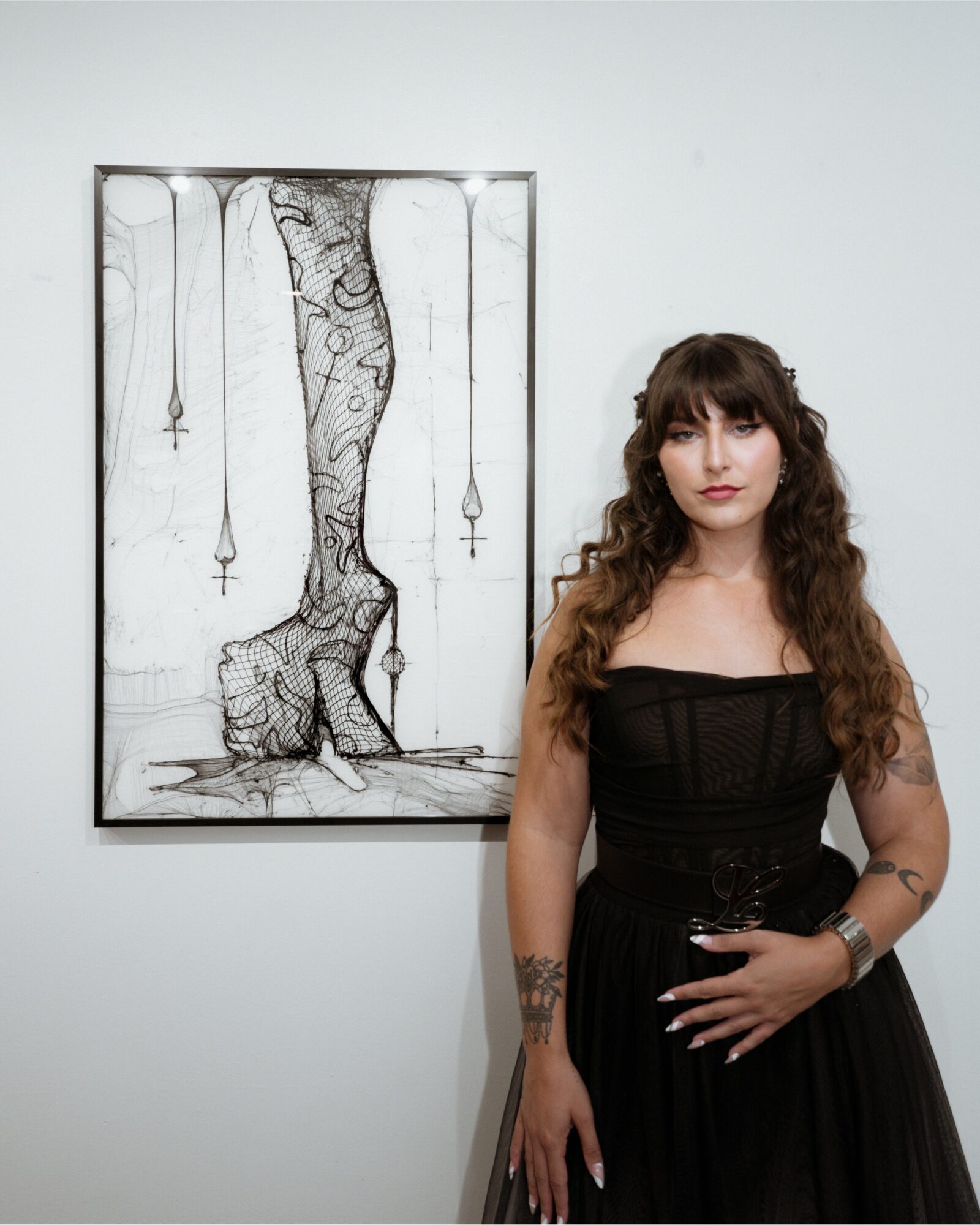 Woman with long curly hair in a black dress standing next to a framed black and white drawing of a tree and a bear, on a white wall.