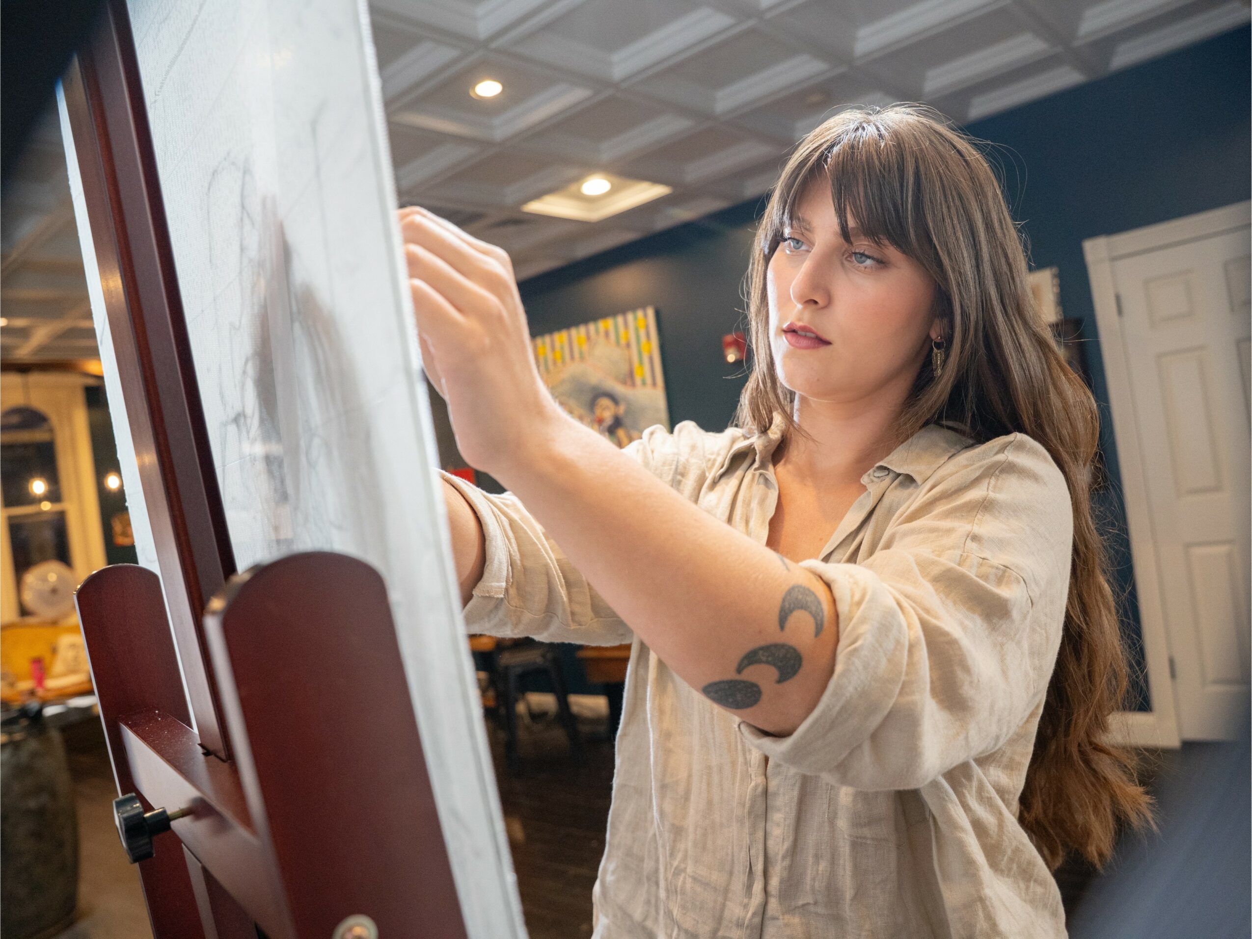 Young woman with long brown hair and a tattoo on her arm, drawing or writing on a whiteboard in a room.