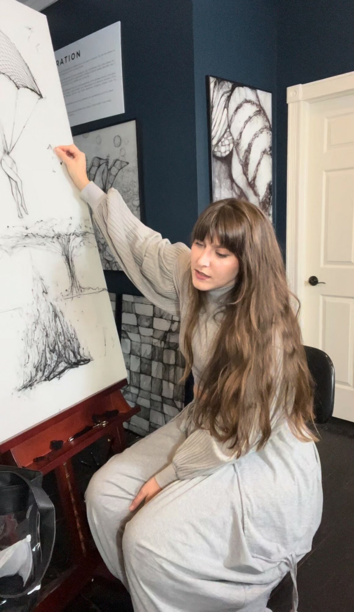 Young woman with long hair drawing on a large canvas in a room with dark blue walls and artwork.