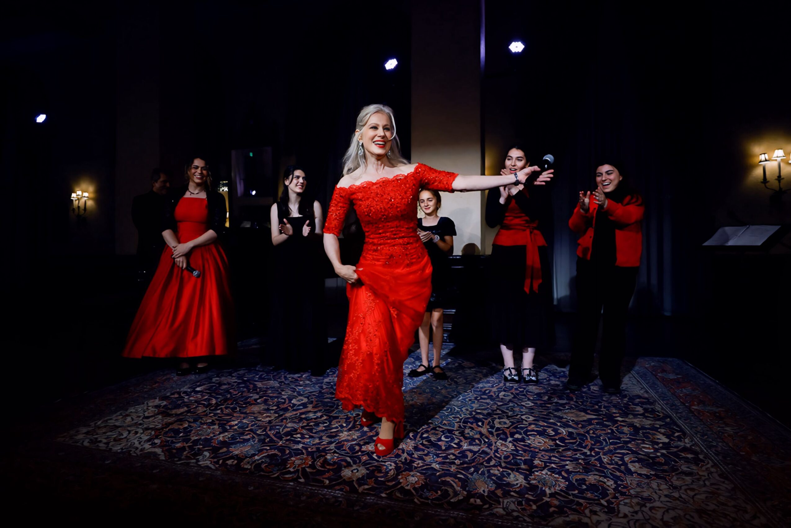 A woman in a red dress walks on stage with five people in the background, all in formal attire, on a dark stage with lighting.