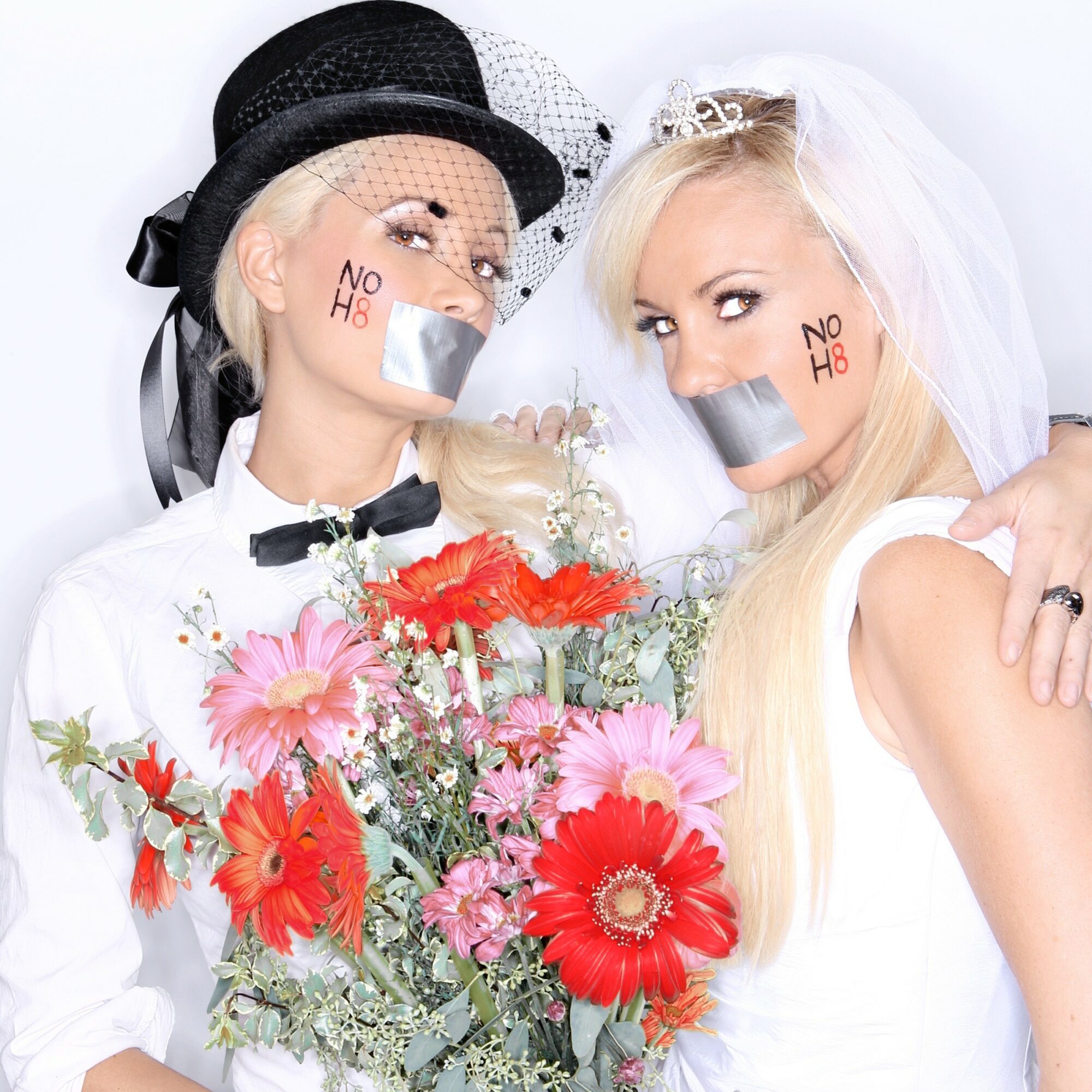 Two women with wedding attire and veils hold a large bouquet of pink and red flowers, with tape over their mouths and 'NO H8' written on their faces.