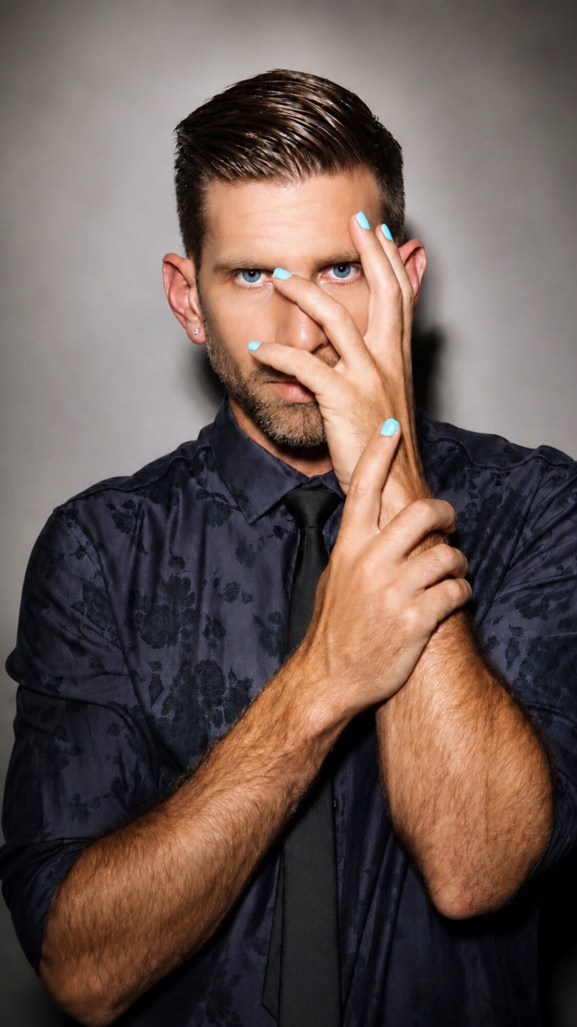Man with dark hair and beard, wearing a dark shirt, holding his hand with painted nails near his face, against a gray background.