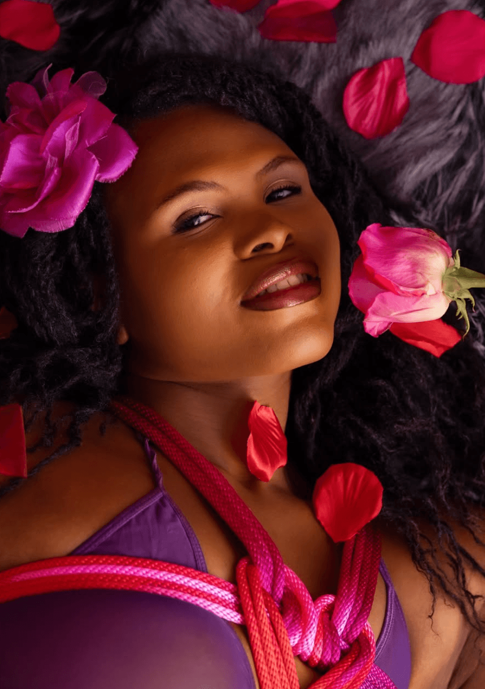 Woman with dark curly hair surrounded by pink and red flower petals, lying on a dark surface, smiling softly.
