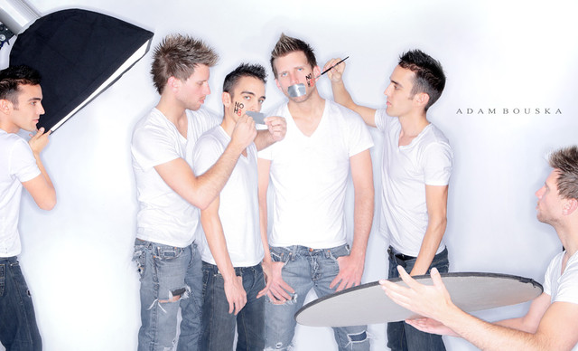 Group of young men in white shirts gathered around a photographer, posing and interacting, in a studio setting.