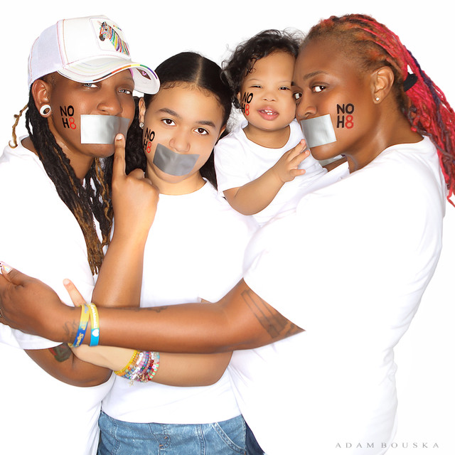 Group of four children with tape over mouths, smiling and hugging, wearing white shirts, against a white background.