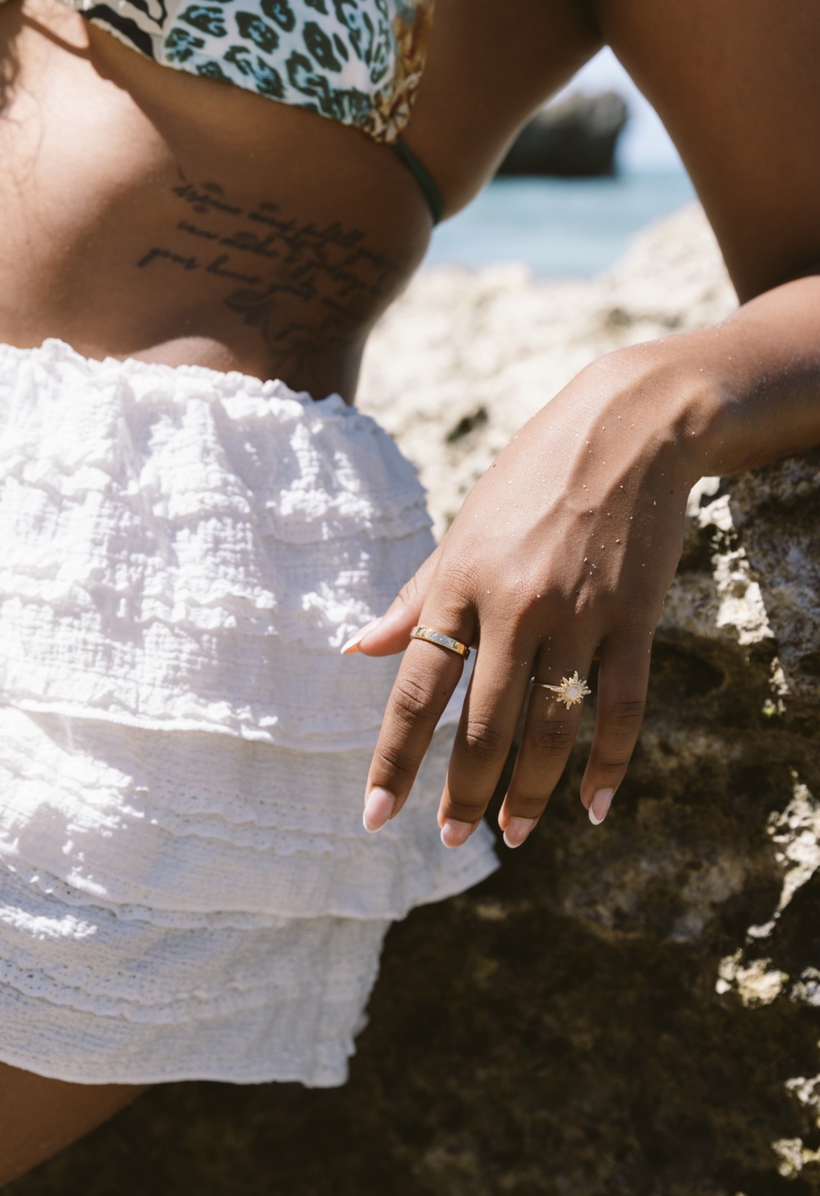 Close-up of a person's hand with rings resting on a rock, wearing a white textured skirt and a patterned top.