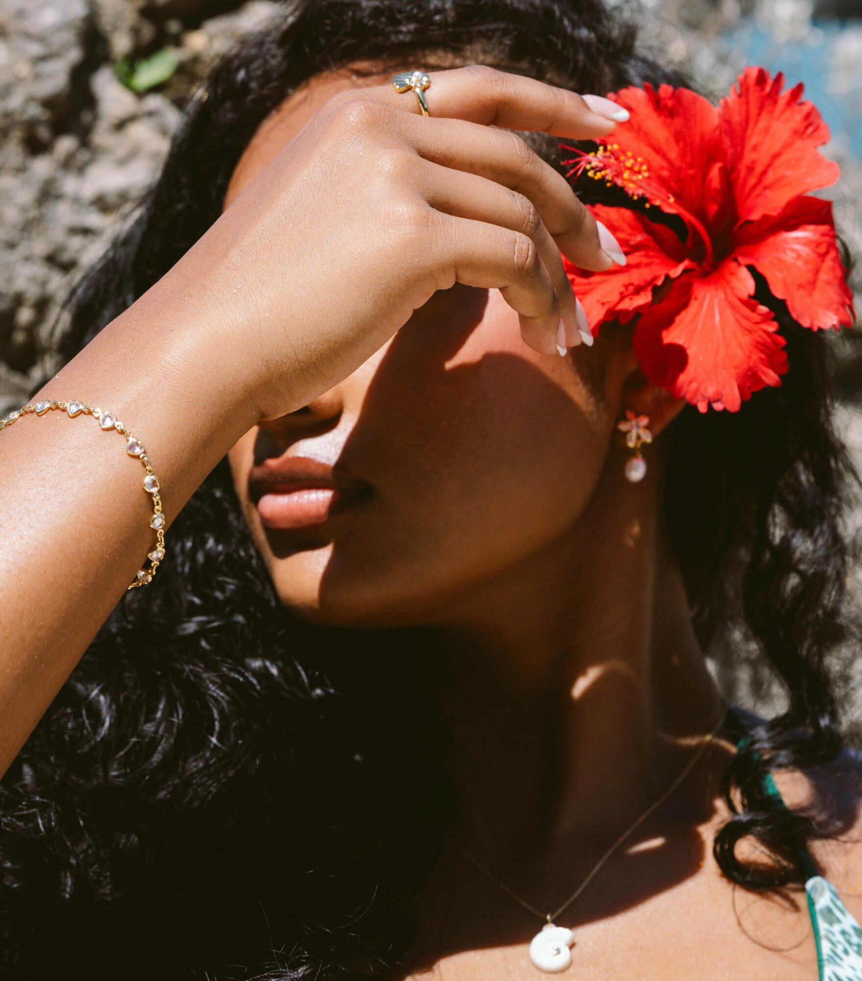 Woman holding a red hibiscus flower near her face, with sunlight on her skin and dark curly hair, outdoors.