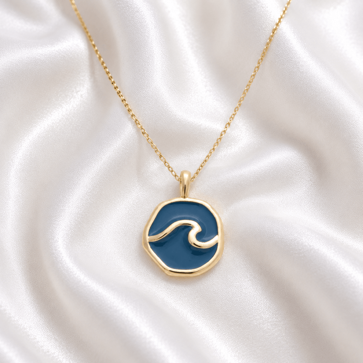 Gold necklace with a round pendant featuring a wave design on a silky white fabric background.