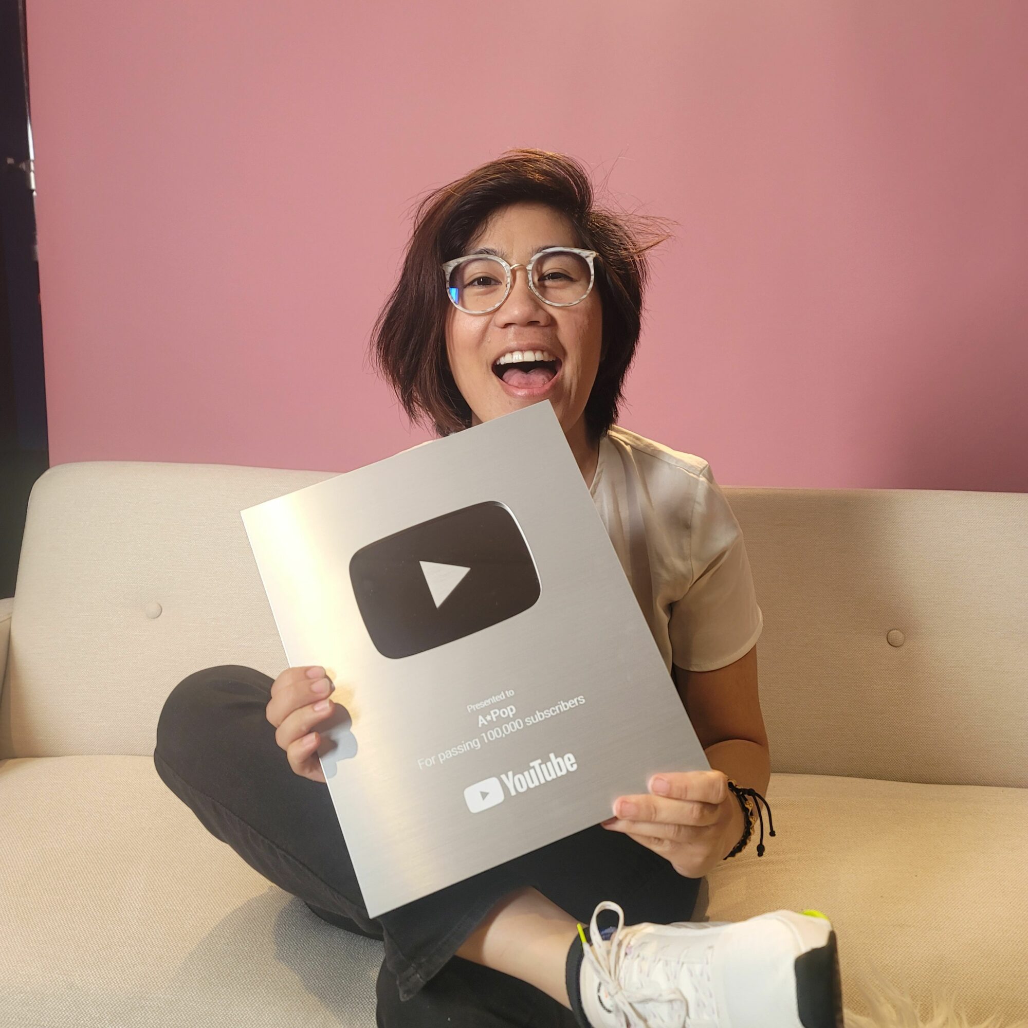 Person with glasses holding a YouTube award plaque, smiling, sitting on a beige couch against a pink wall.