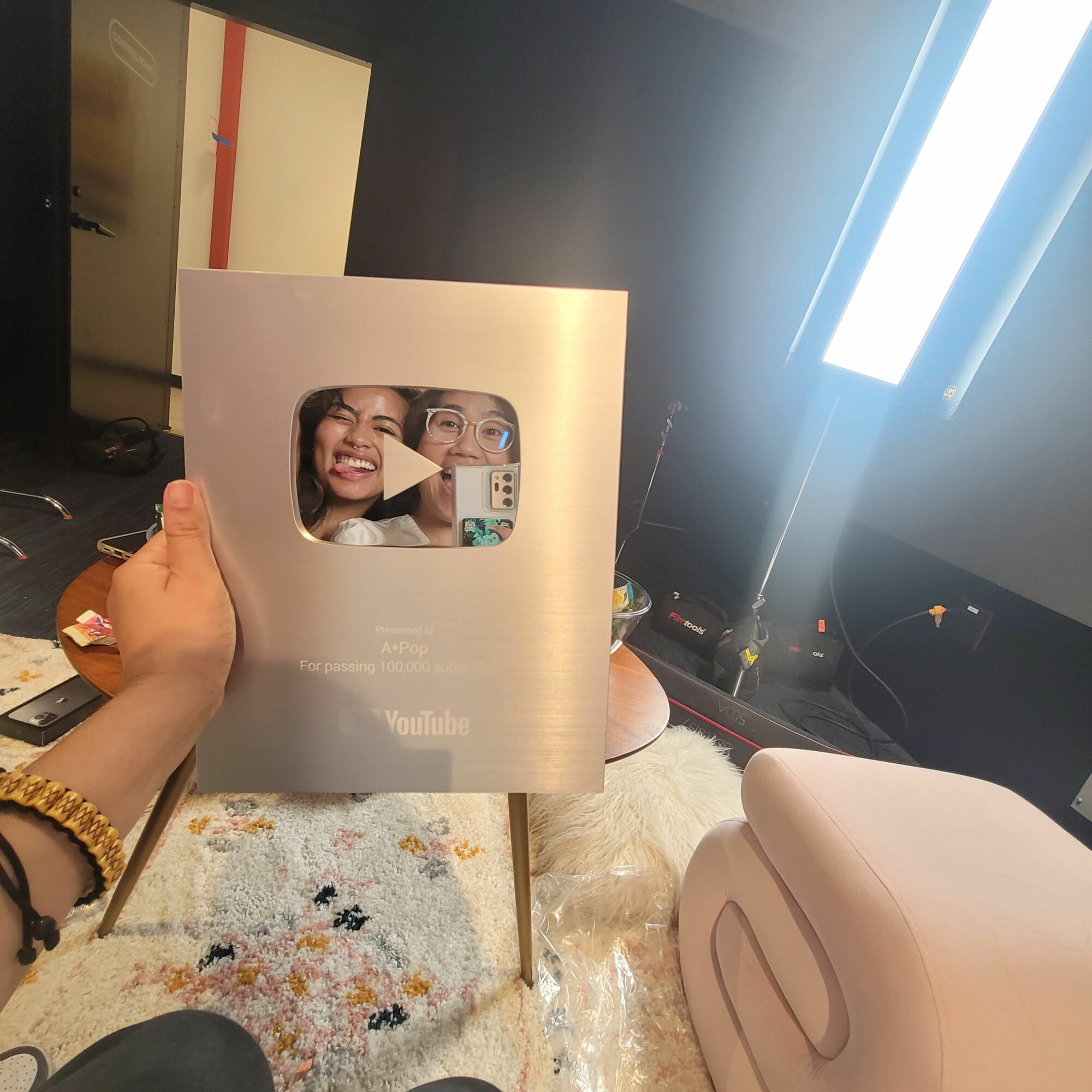 Person holding a YouTube Silver Play Button award with a play icon, in a room with a beige chair and a colorful rug.