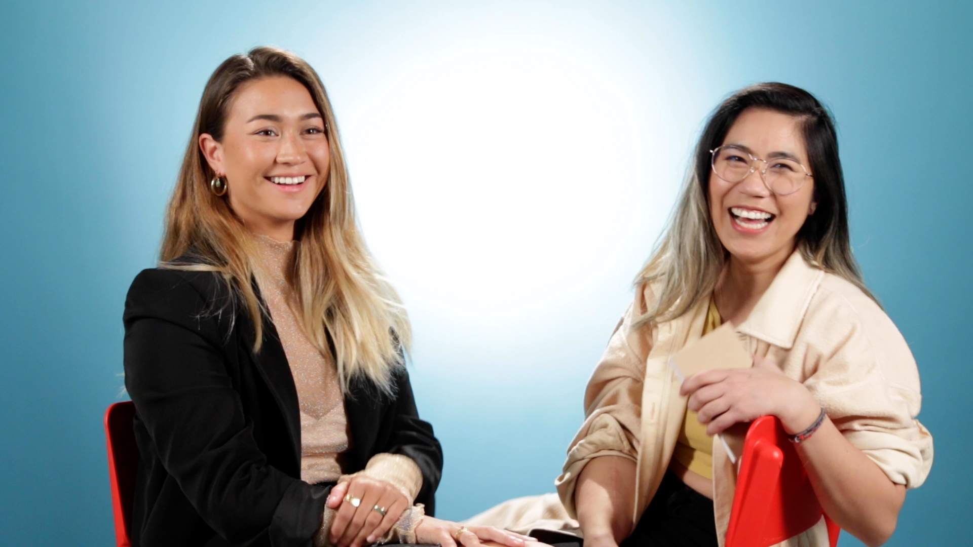Two women sitting and smiling, one with long blonde hair and the other with long dark hair, against a blue background.