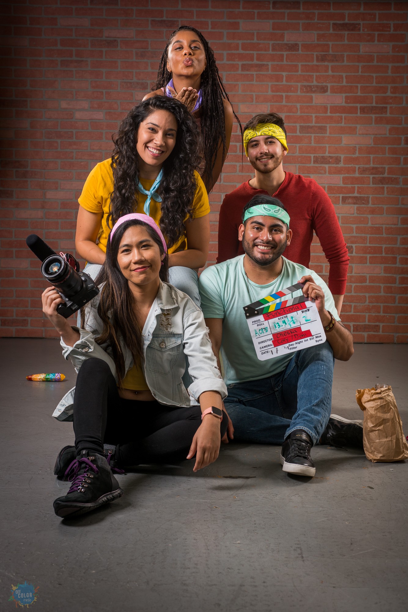 Five diverse people, three women and two men, pose with a brick wall background, holding a camera and a clapperboard, smiling.
