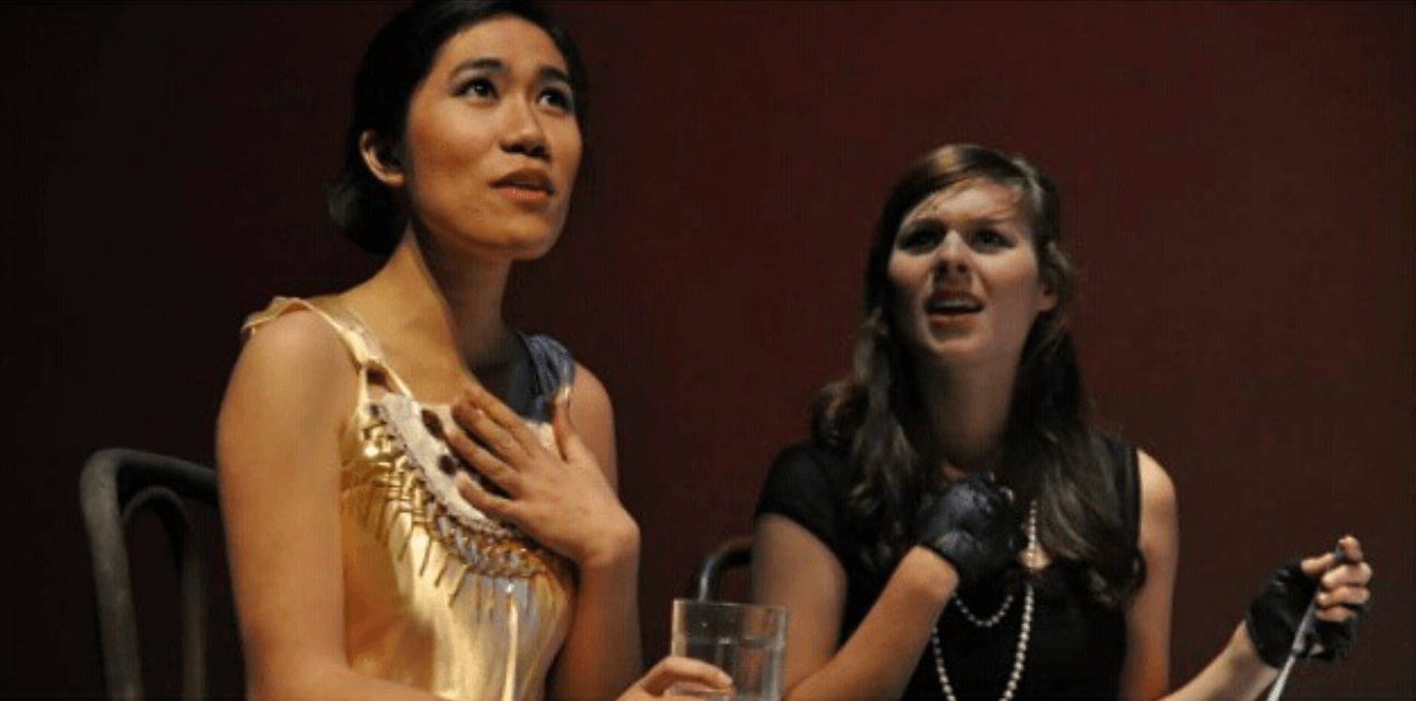 Two women sitting, one with hand on chest, the other smiling, against a dark background.