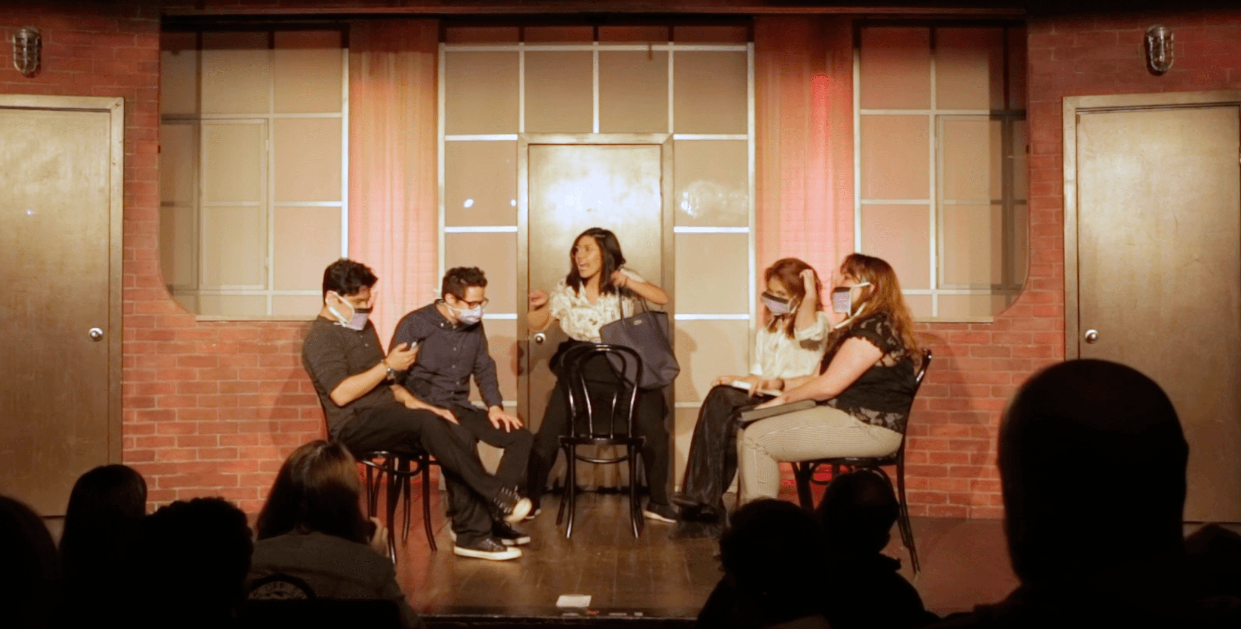 Five people sit on stage in front of a brick wall with large windows, engaging in a discussion or performance.