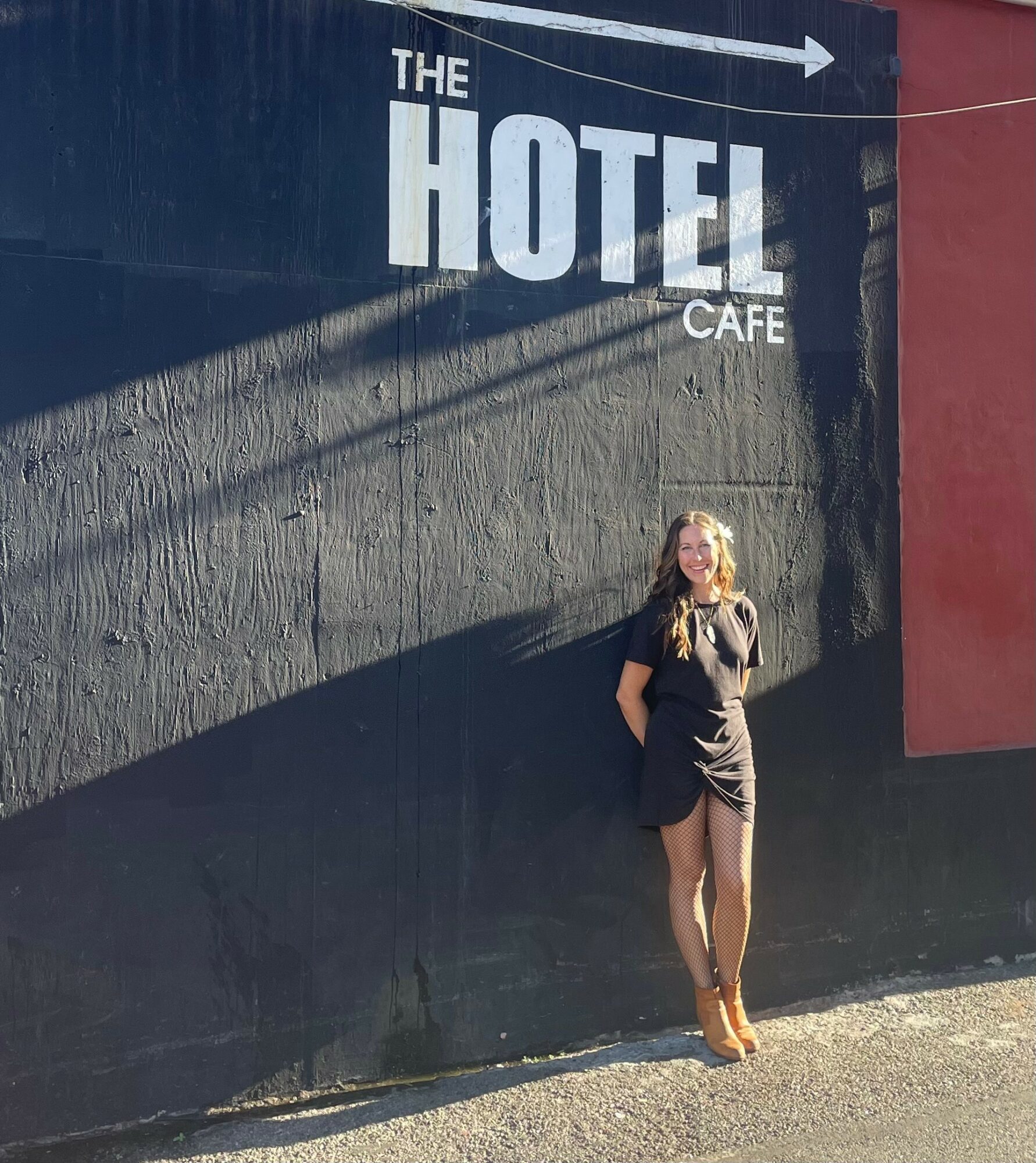 Woman standing against a black wall with a sign reading 'The Hotel Cafe' in white letters, sunlight casting shadows.