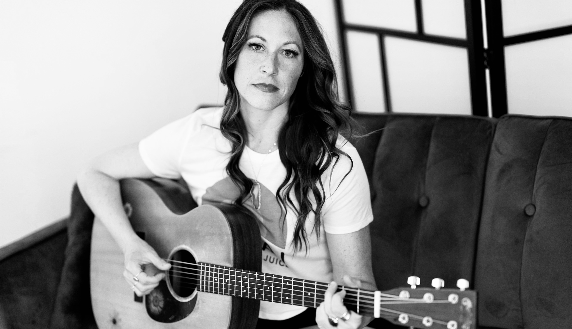 Woman with long hair playing an acoustic guitar on a sofa, black and white photo.