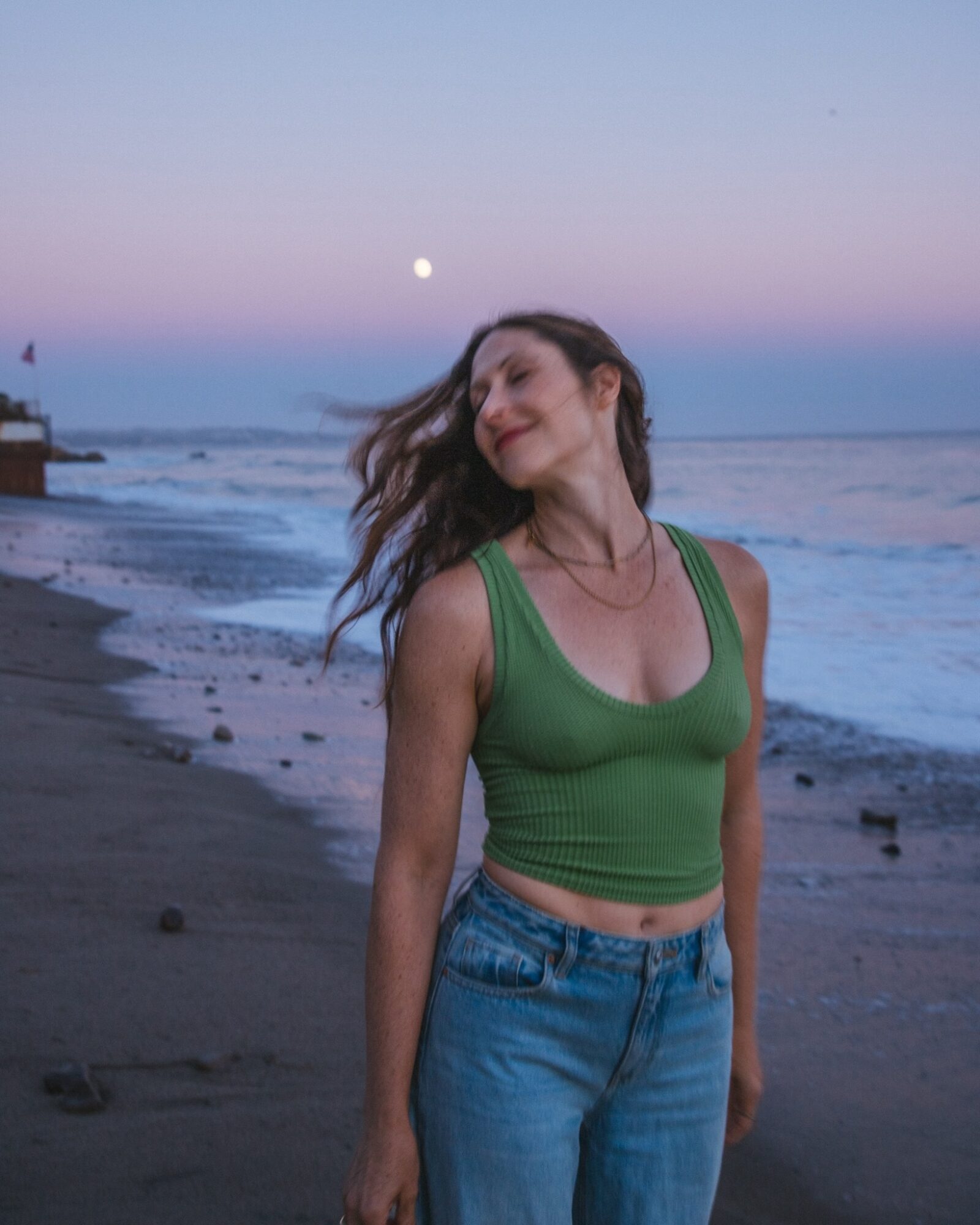 Woman with long hair in a green tank top and jeans standing on a beach at sunset, smiling with eyes closed.