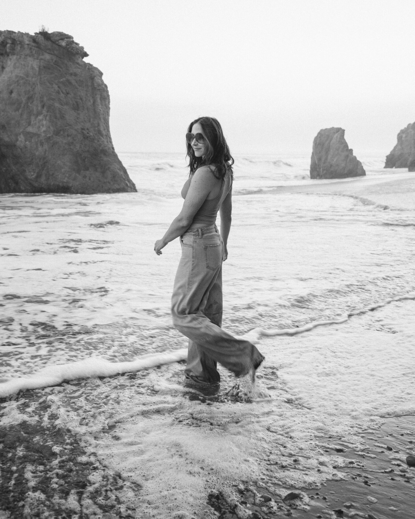Woman standing in shallow ocean water with large rocks in background, facing sideways, smiling, wearing pants and top.