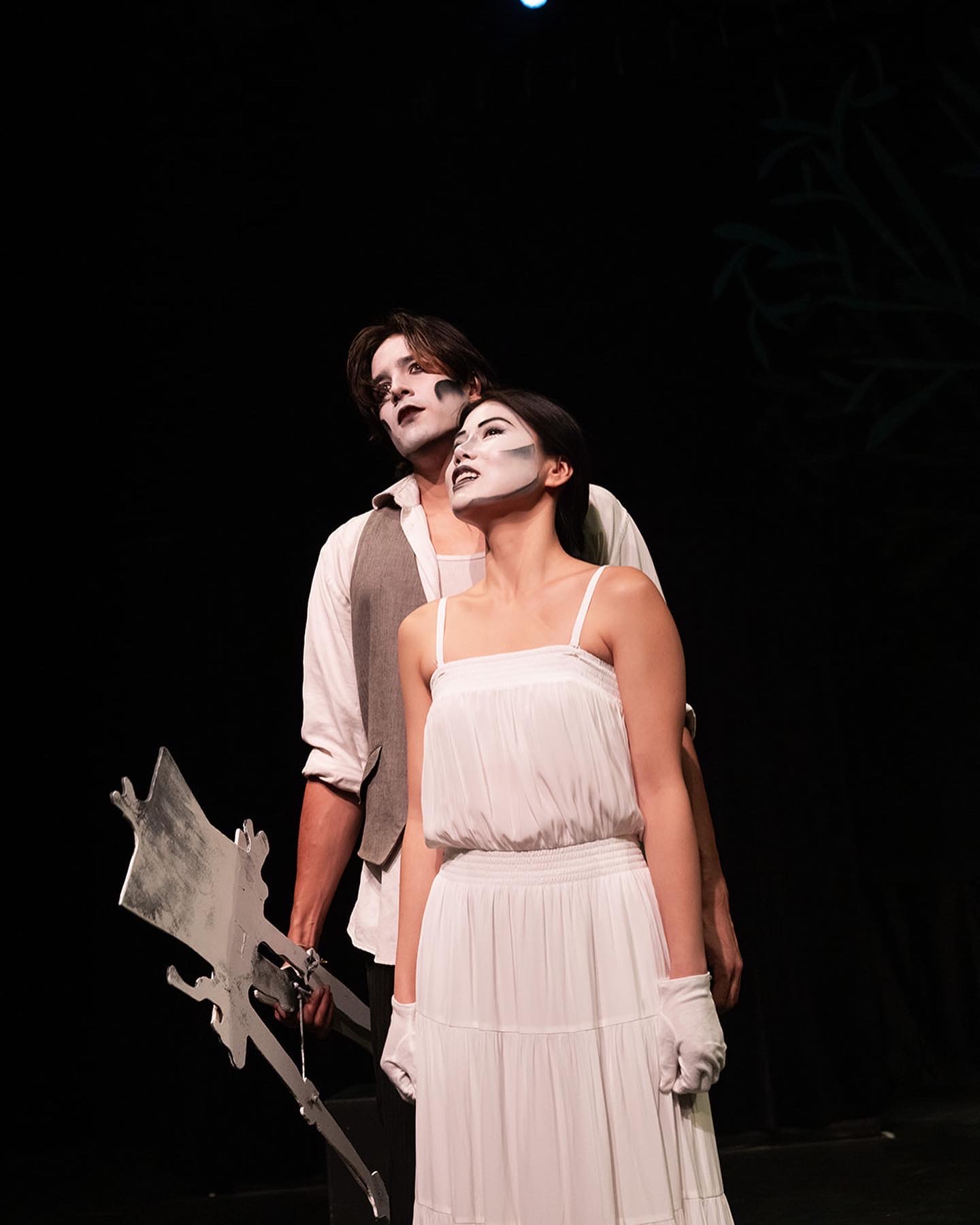 Two performers on stage, one with dark hair and makeup, the other with light hair, both dressed in light-colored costumes, standing close together against a dark background.