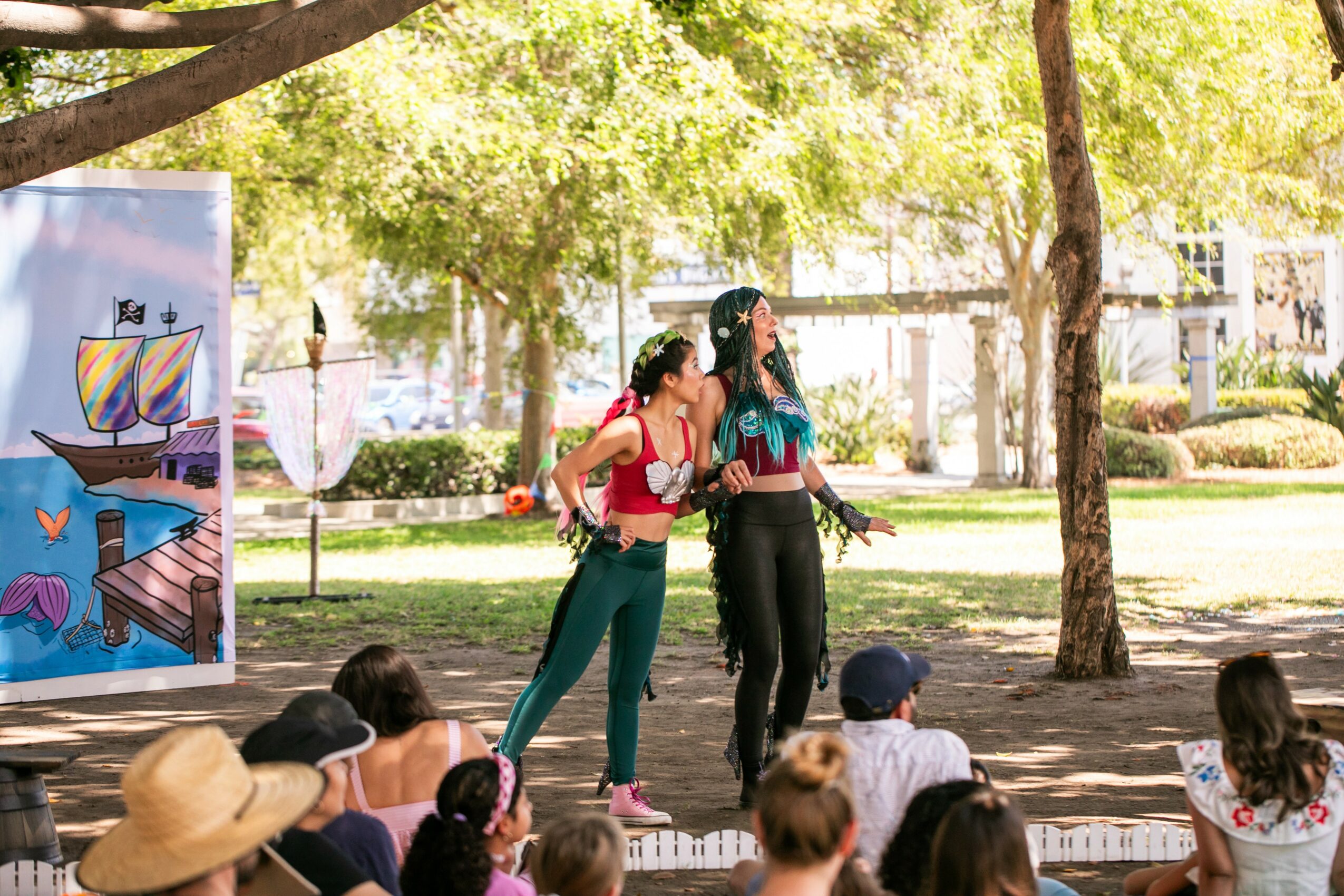 Two women stand on a stage outdoors, speaking to an audience in a park with trees and sunlight.