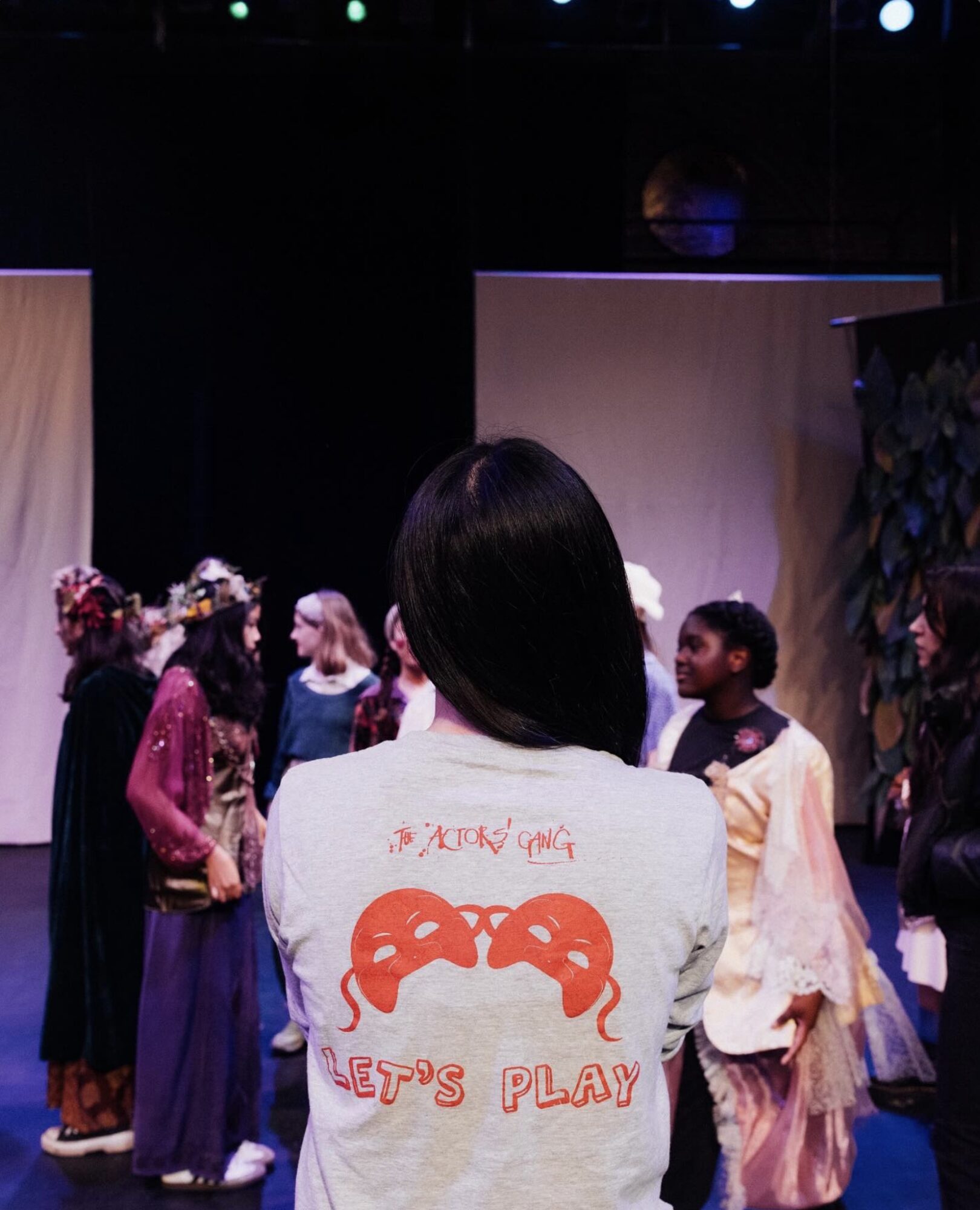 Person with dark hair facing a stage with children performing, stage background with plants and panels, dim lighting.