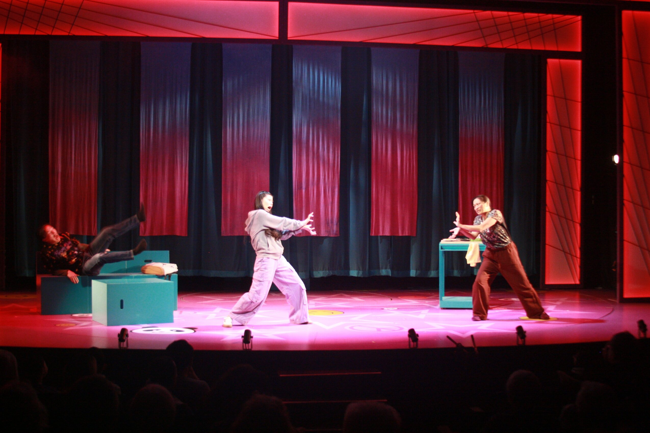 Three performers on stage with colorful curtains, two are dancing and one is lying on a teal box, stage lit with red and blue lights.