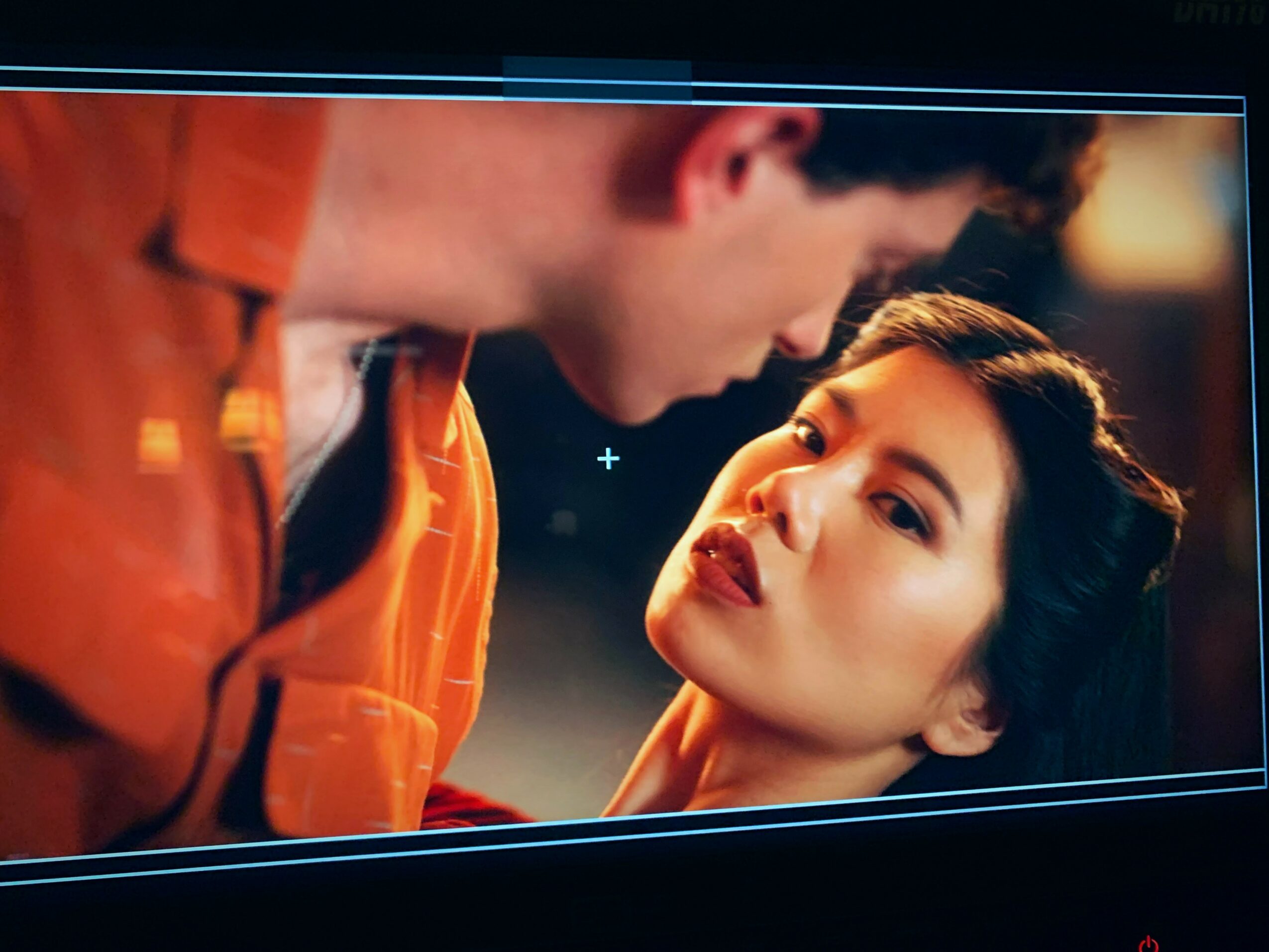 A woman looks at a man with a serious expression, both are close and indoors with warm lighting.