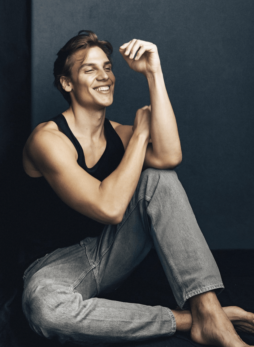 Woman sitting against a dark wall, smiling, wearing a black top and gray pants, with one arm raised and bent.