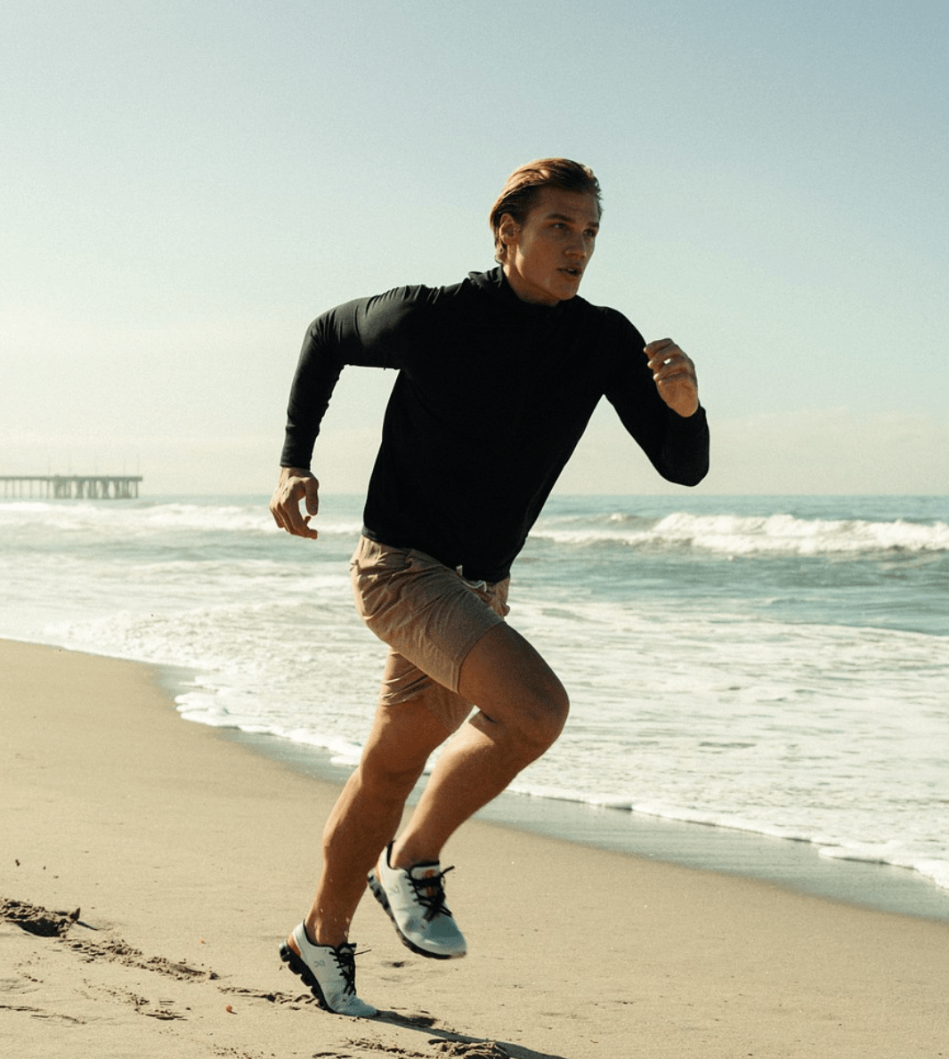 Person running on beach with ocean and sky in background, wearing black top and beige shorts.